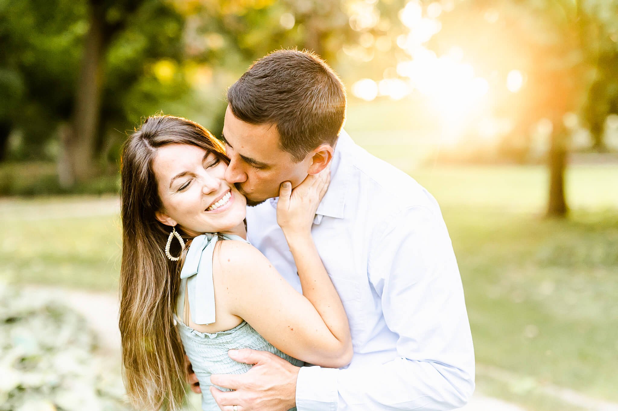 Man kisses girlfriend on the cheek in a blue collared shirt in a green grassy park at sunset