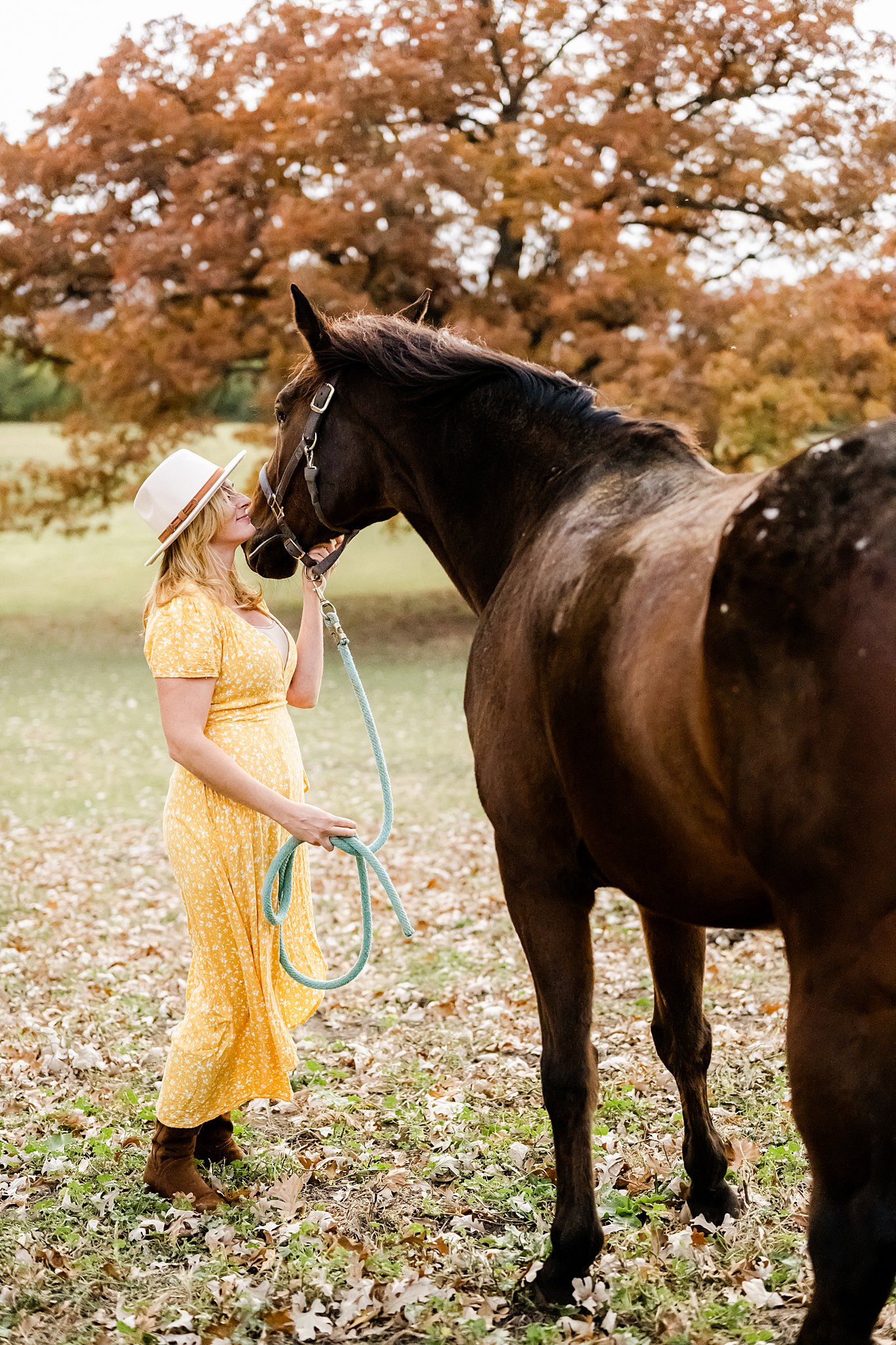 Women in yellow sundress and large hat holds horse lead and scratches the horse's chin in a large open field horseback riding chicago