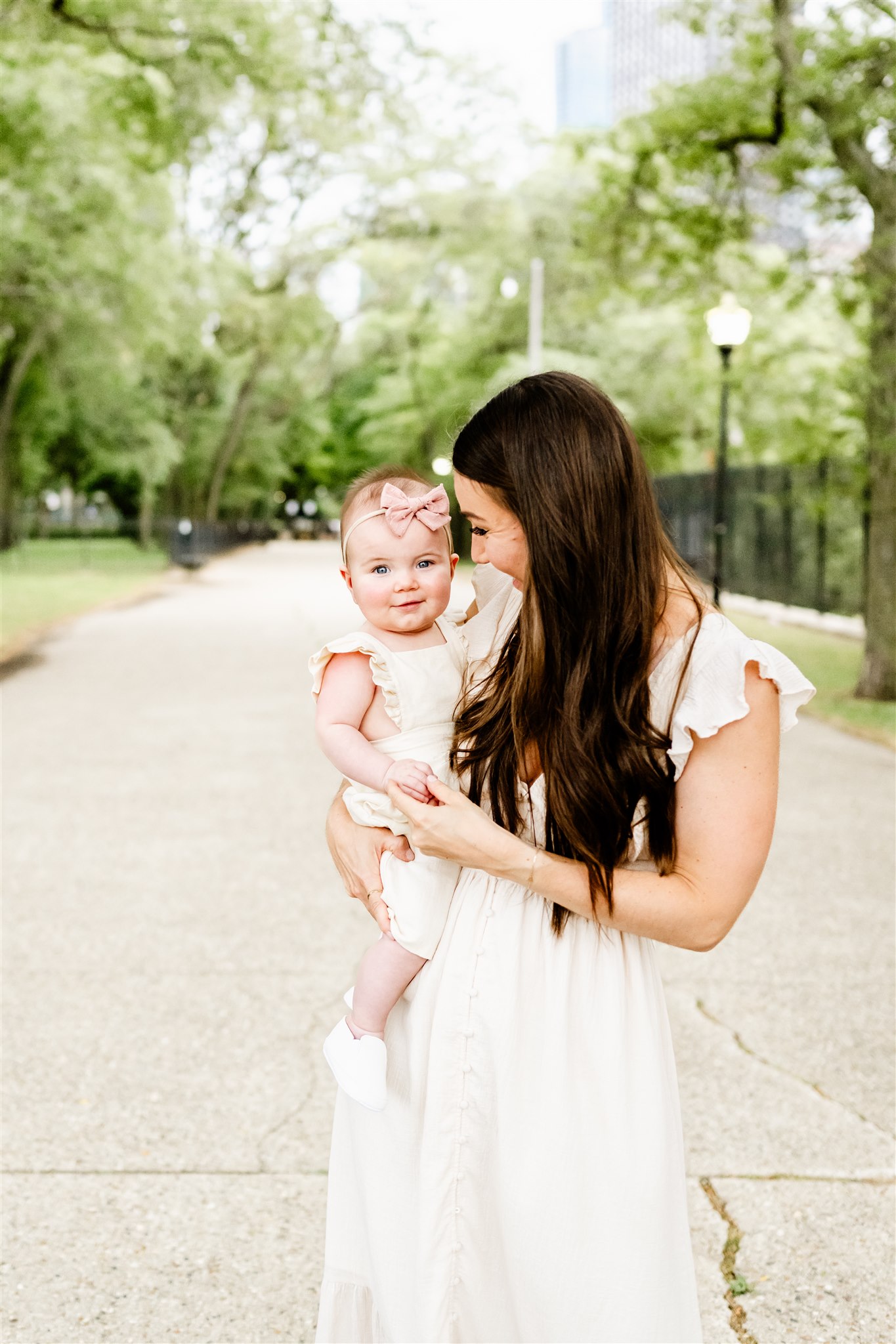 A mother in a white dress walks through a park sidewalk with her infant daughter in a pink bow on her hip