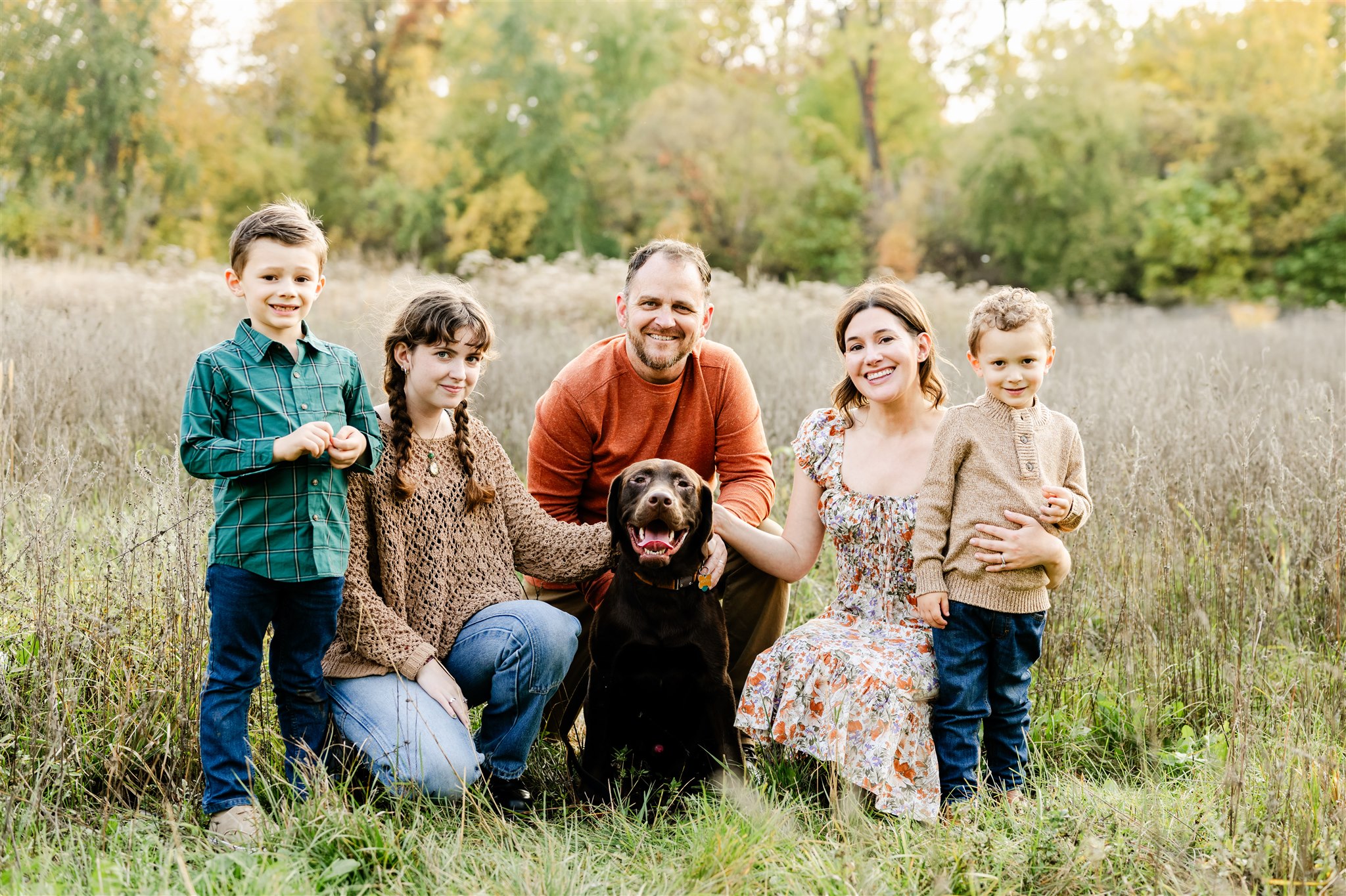 A mom and dad kneel in a field of tall grass with their three children and chocolate lab