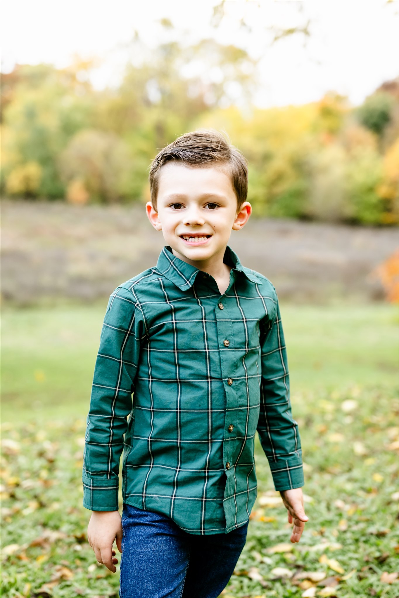 A young boy in a green plaid shirt smiles while standing in a grassy park