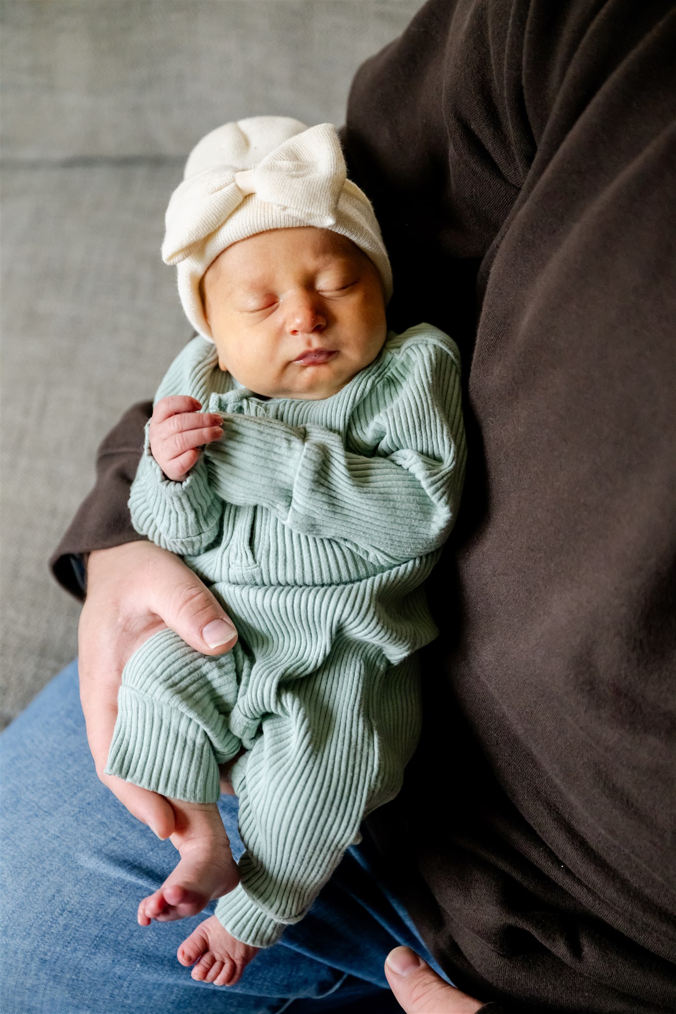 A newborn baby sleeps in dad's arms while wearing a white beanie with a bow and a green onesie