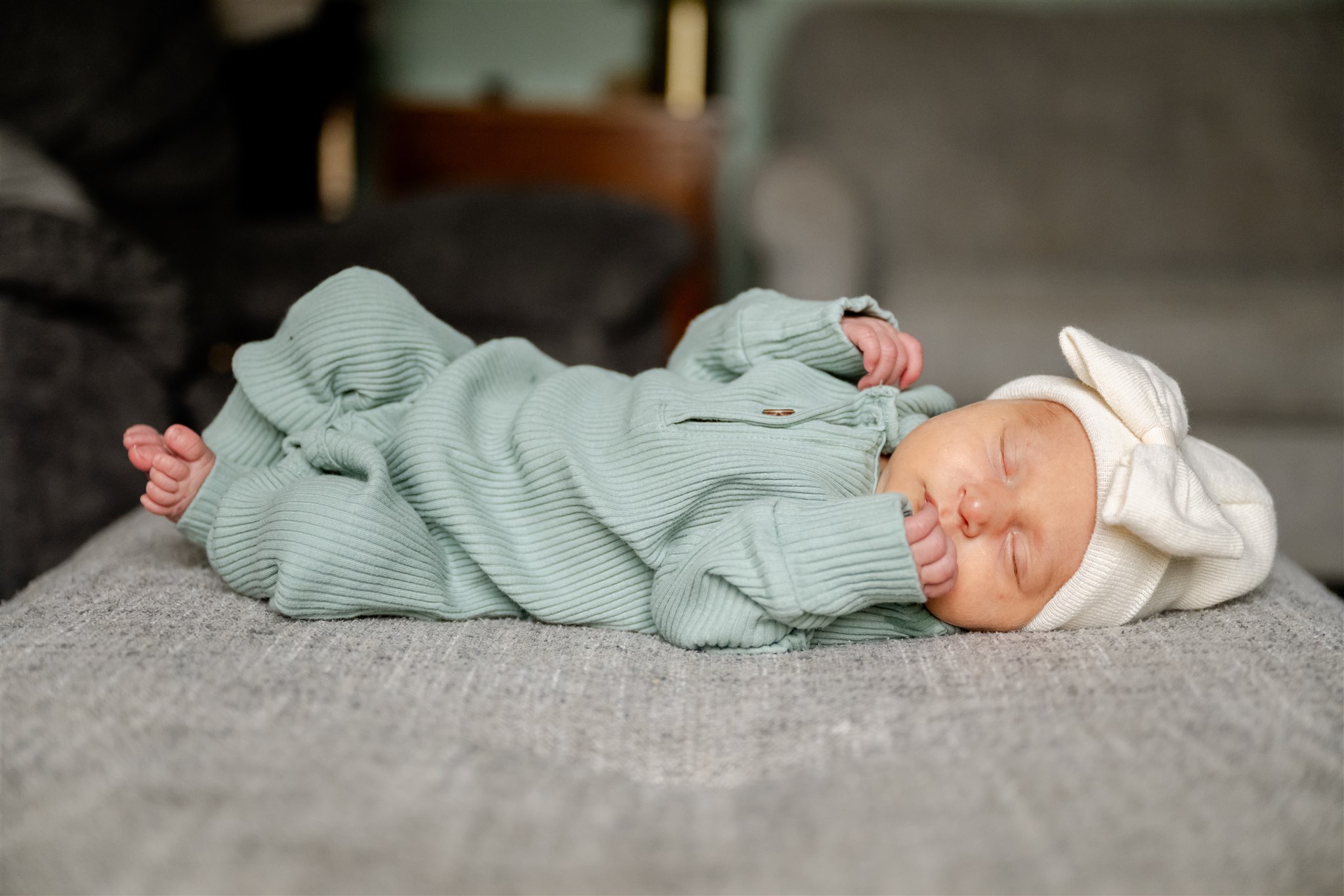 A newborn baby in a green onesie and white beanie with a bow sleeps on a grey couch thanks to Chicago Baby Stores