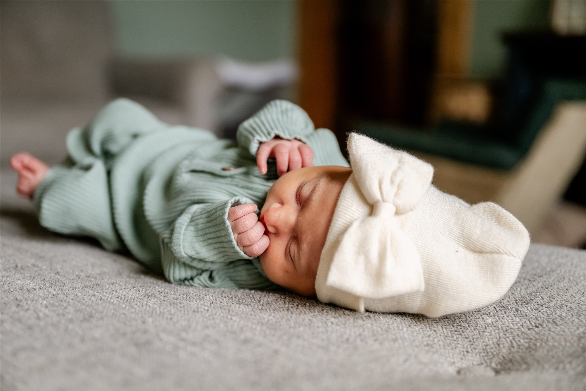 A newborn baby sleeps on a couch in a green onesie and large white beanie from Chicago Baby Stores