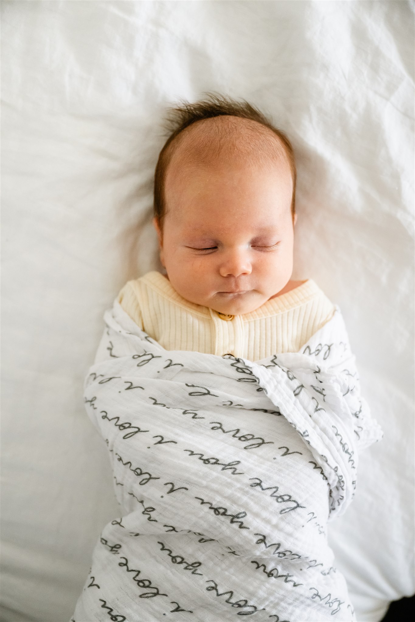 A newborn baby sleeps in a text covered swaddle on a white bed