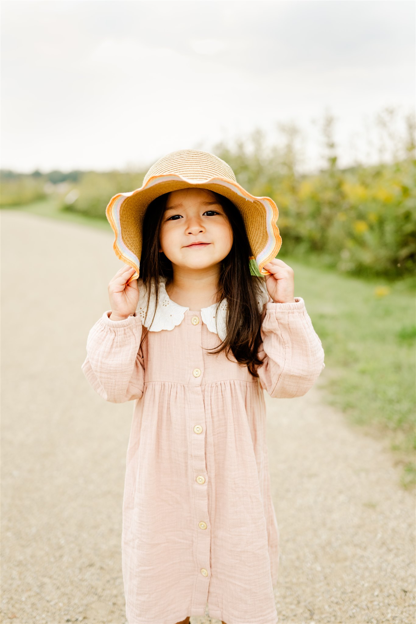 A young toddler girl in a pink dress plays in a park path with a large yellow sun hat