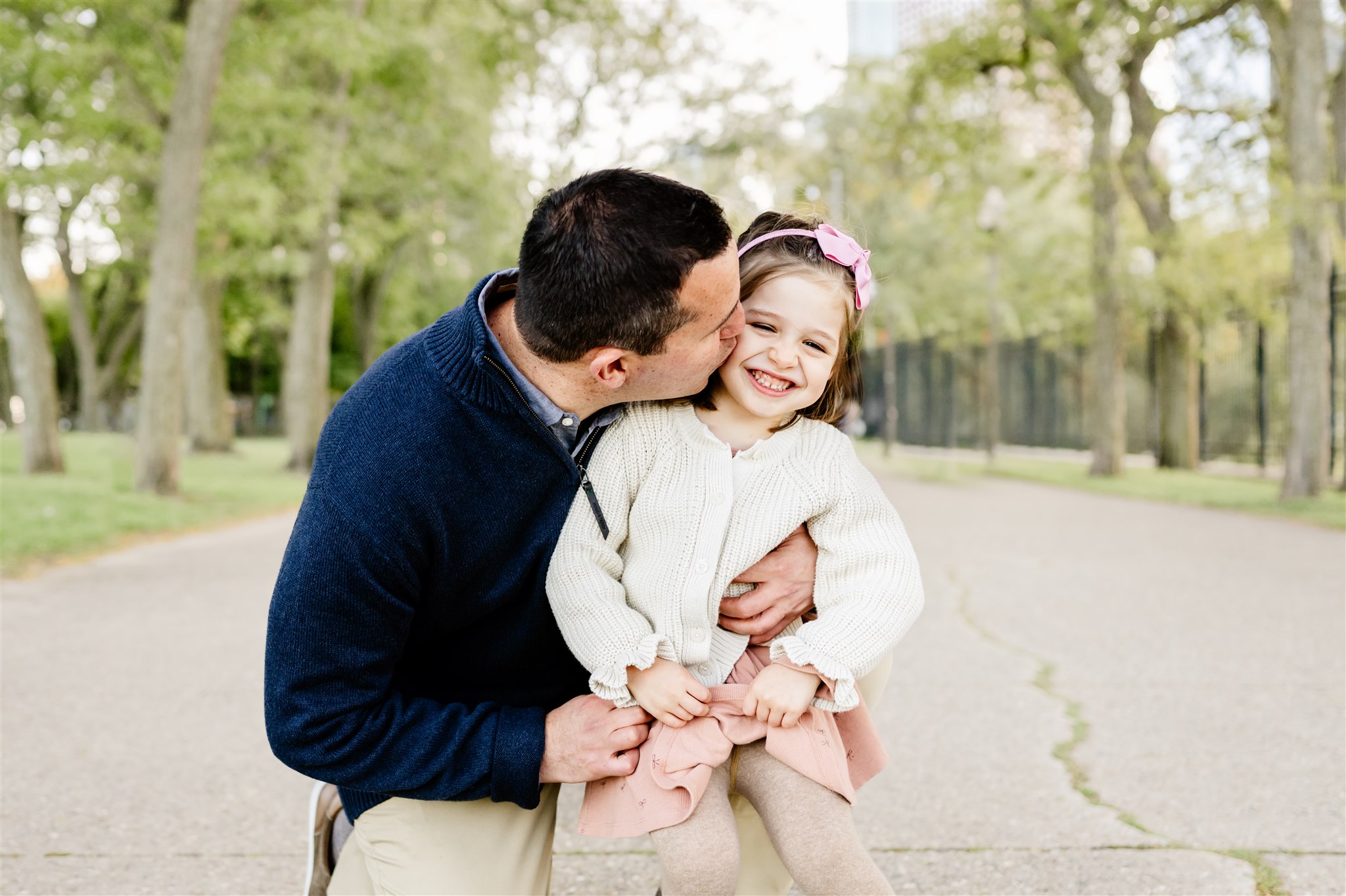 A dad in a blue sweater kisses the cheek of his smiling toddler daughter in a park sidewalk