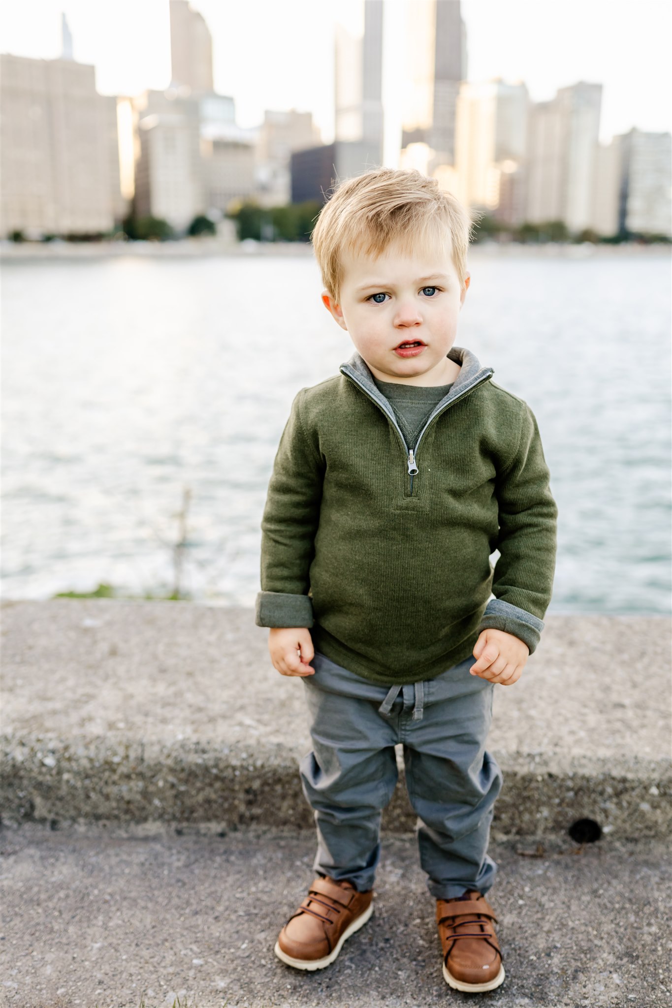 A toddler boy in a green sweater stands in a downtown waterfront park before Ice Skating Chicago