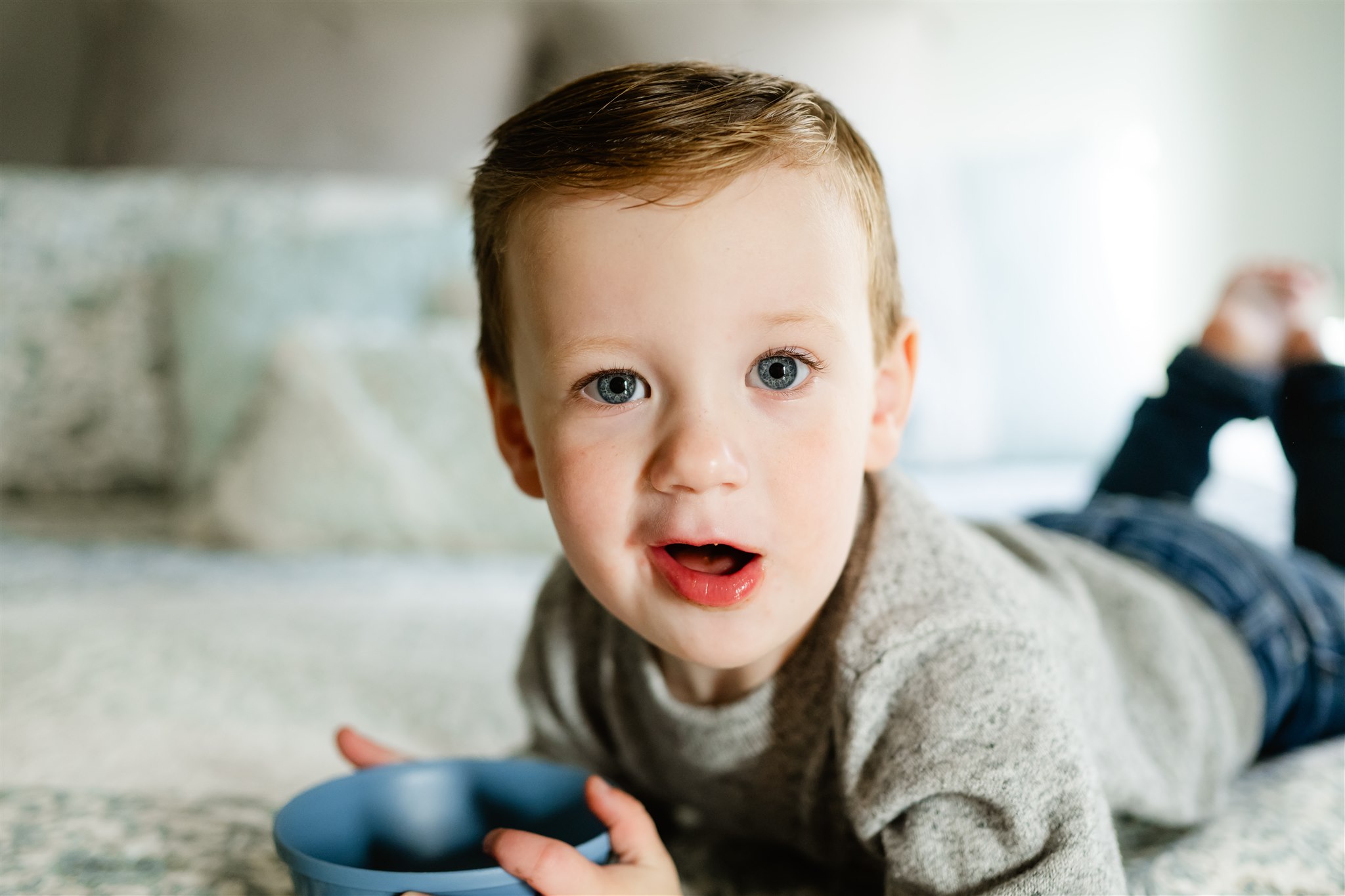 A toddler in a grey sweater lays on a bed eating a bowl of snacks