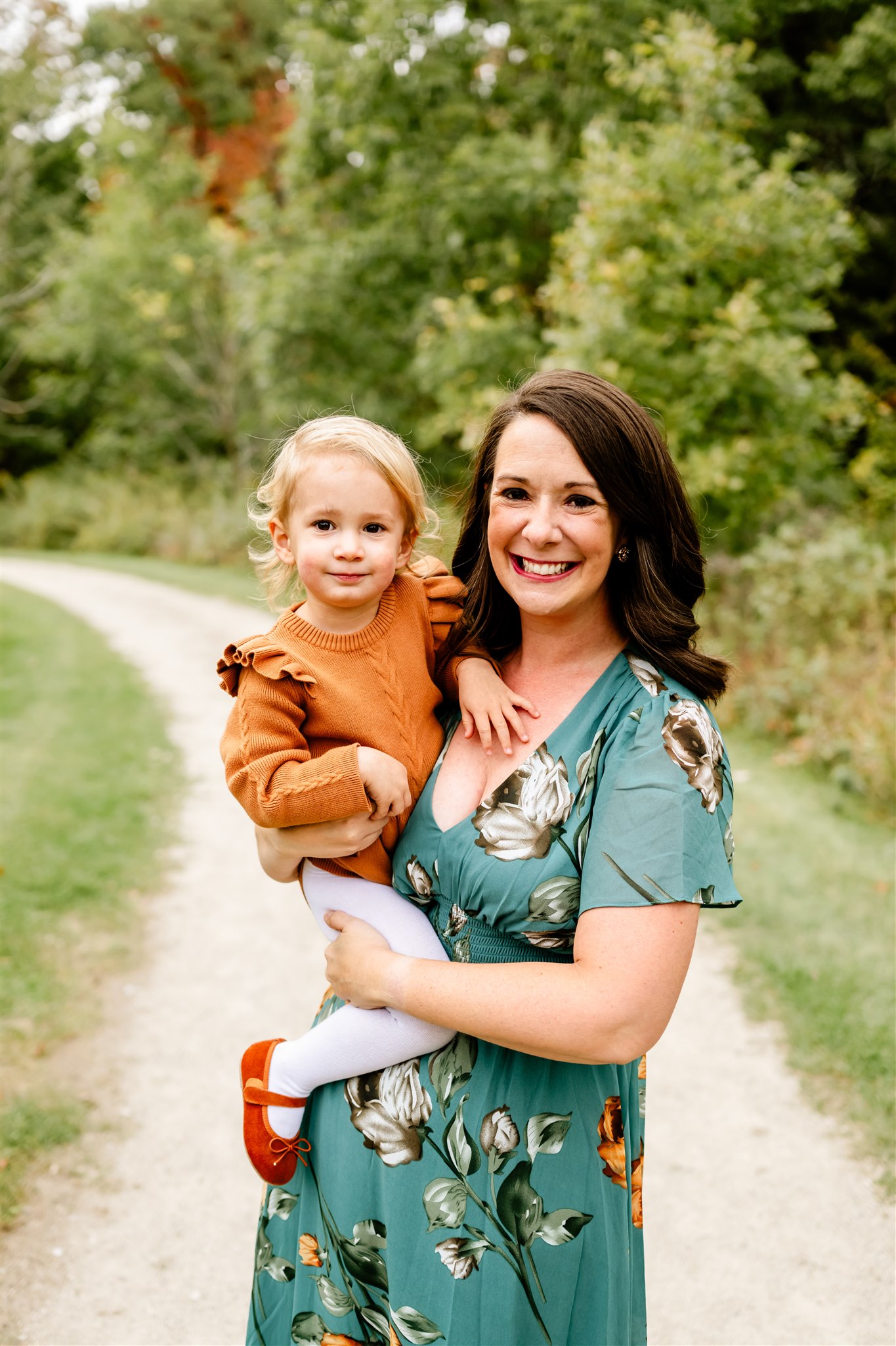 A mother in a teal dress holds her daughter in an orange dress on her hip while walking down a park trail after visiting Kid Birthday Party Ideas Chicago