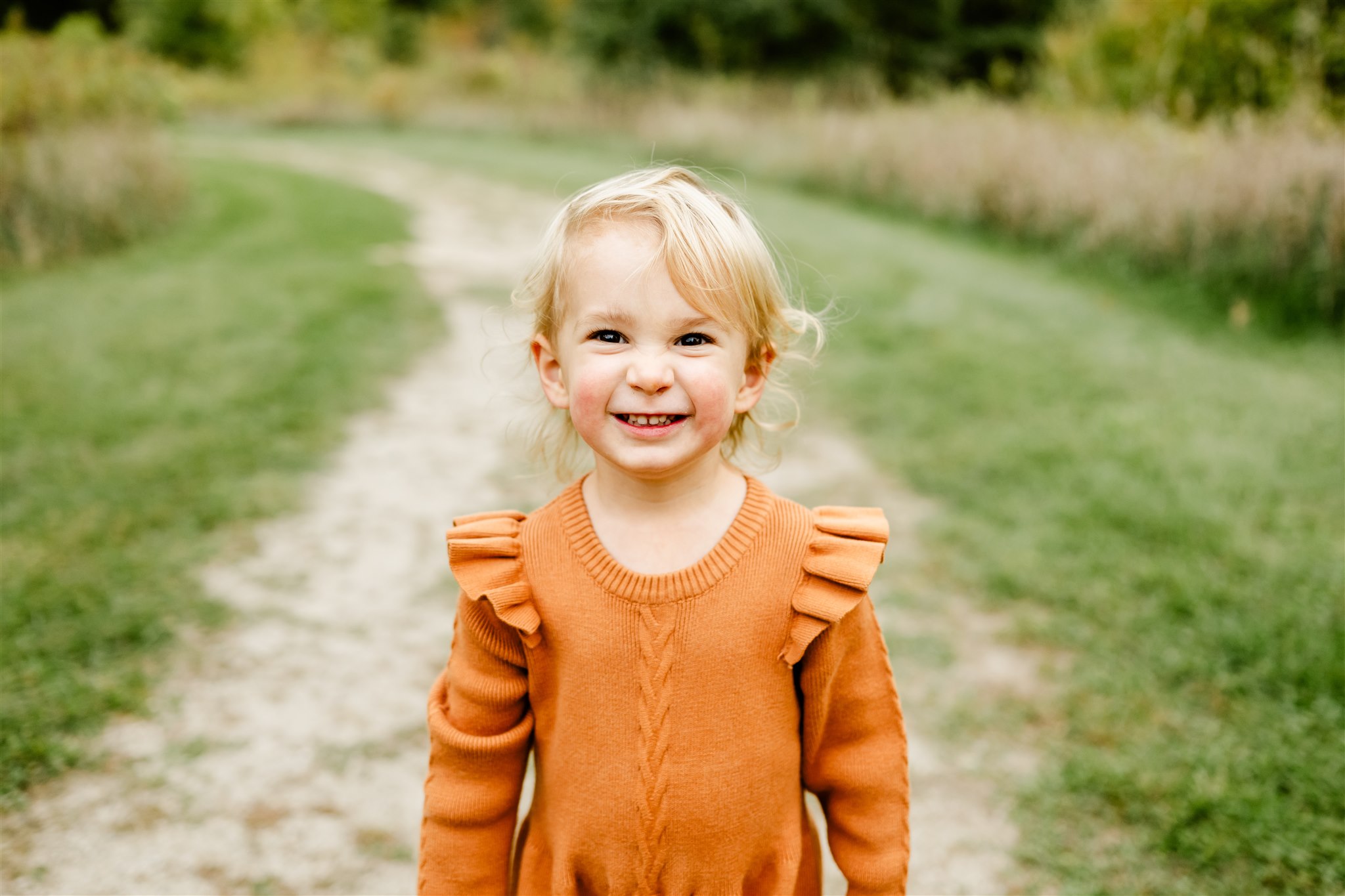 A toddler girl in an orange dress smiles large while standing in a park trail after talking about Kid Birthday Party Ideas Chicago