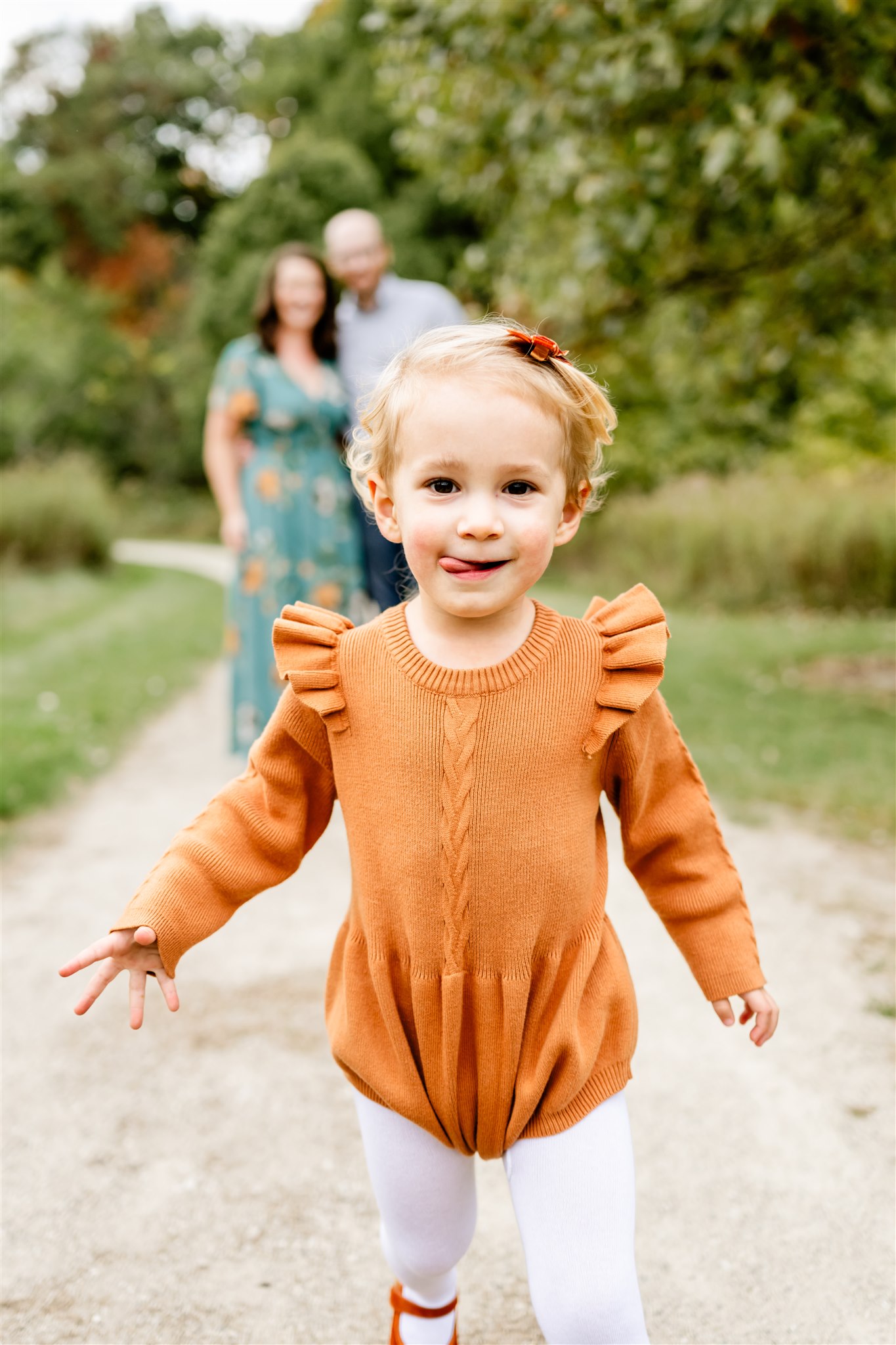 A young toddler in an orange dress walks down a park trail away from mom and dad