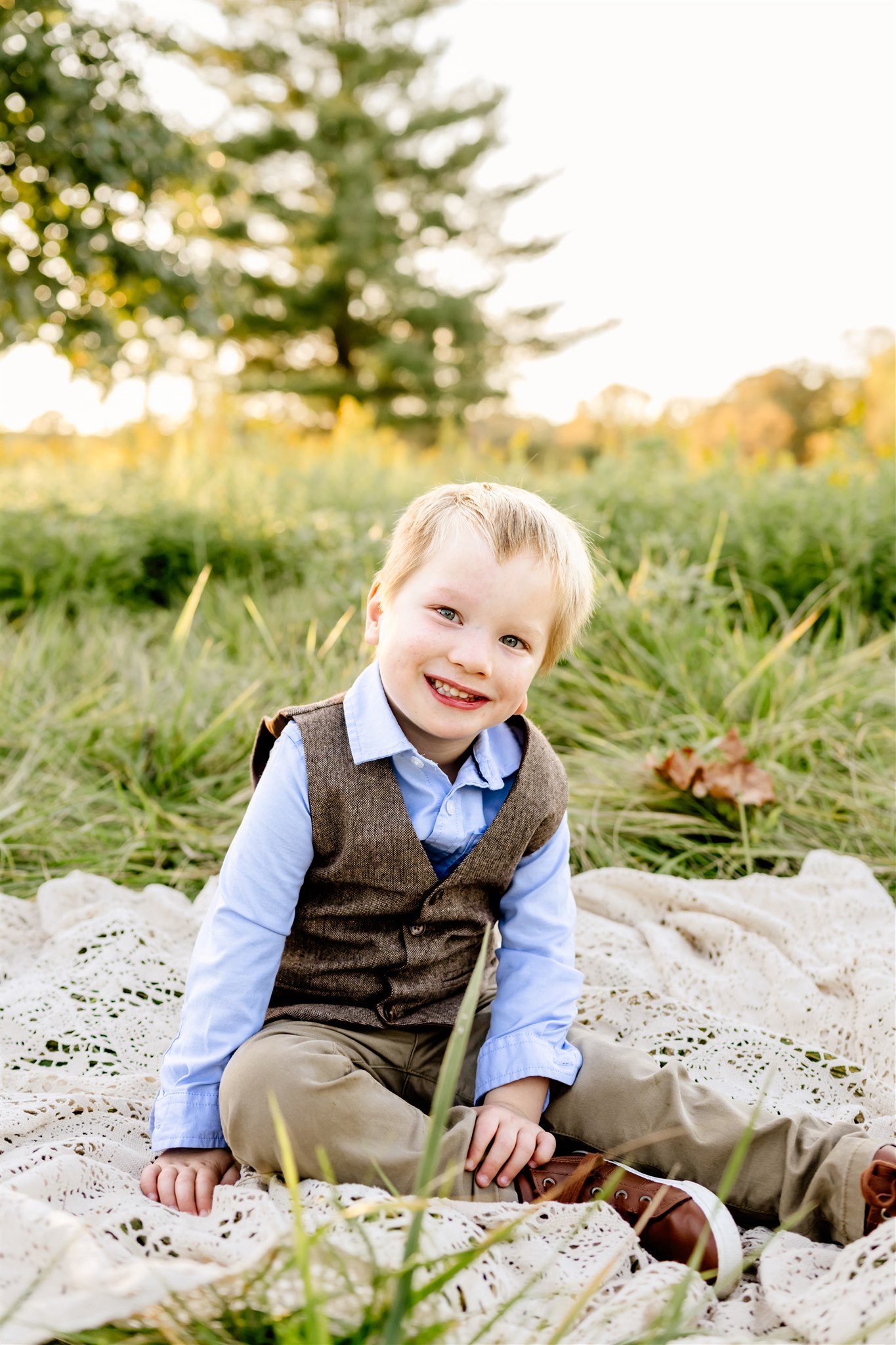 A young boy in a brown vest and blue shirt sits on a picnic blanket in a park at sunset
