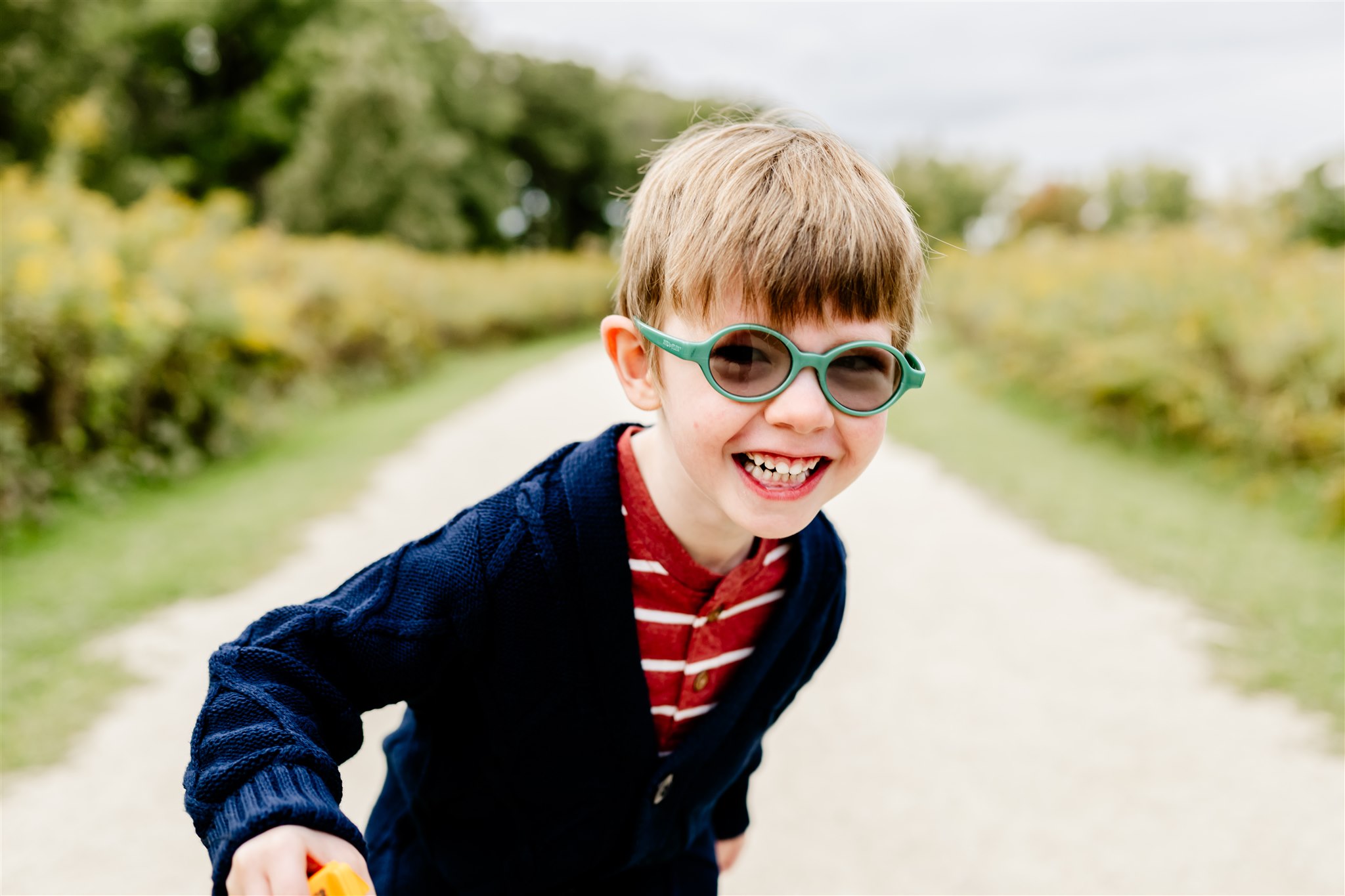 A young boy laughs while walking through a park path in a blue sweater and green glasses
