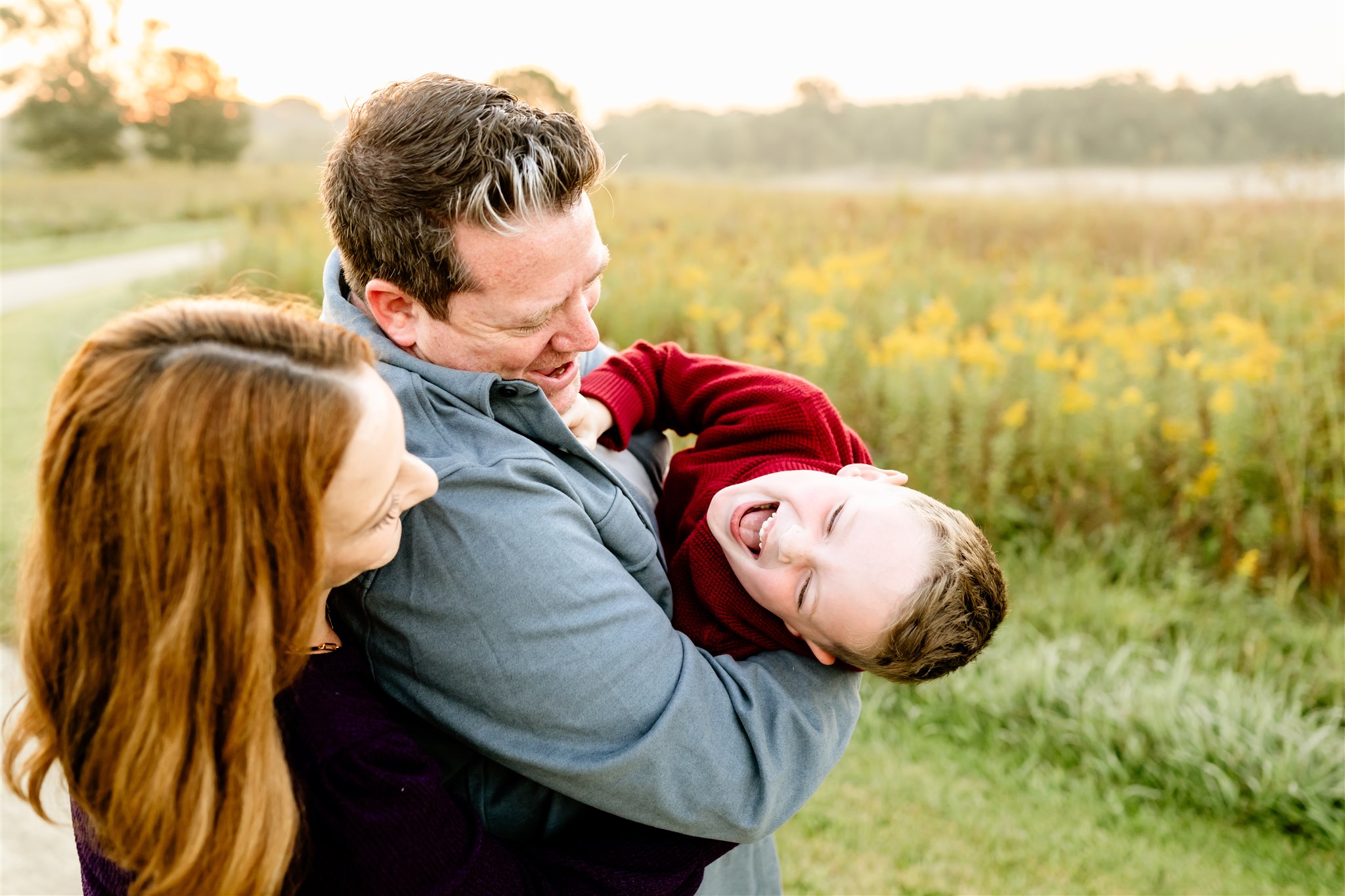 A mom and dad play with their ticklish son in a park path through wildflowers at sunset after visiting Montessori Schools in Chicago Suburbs