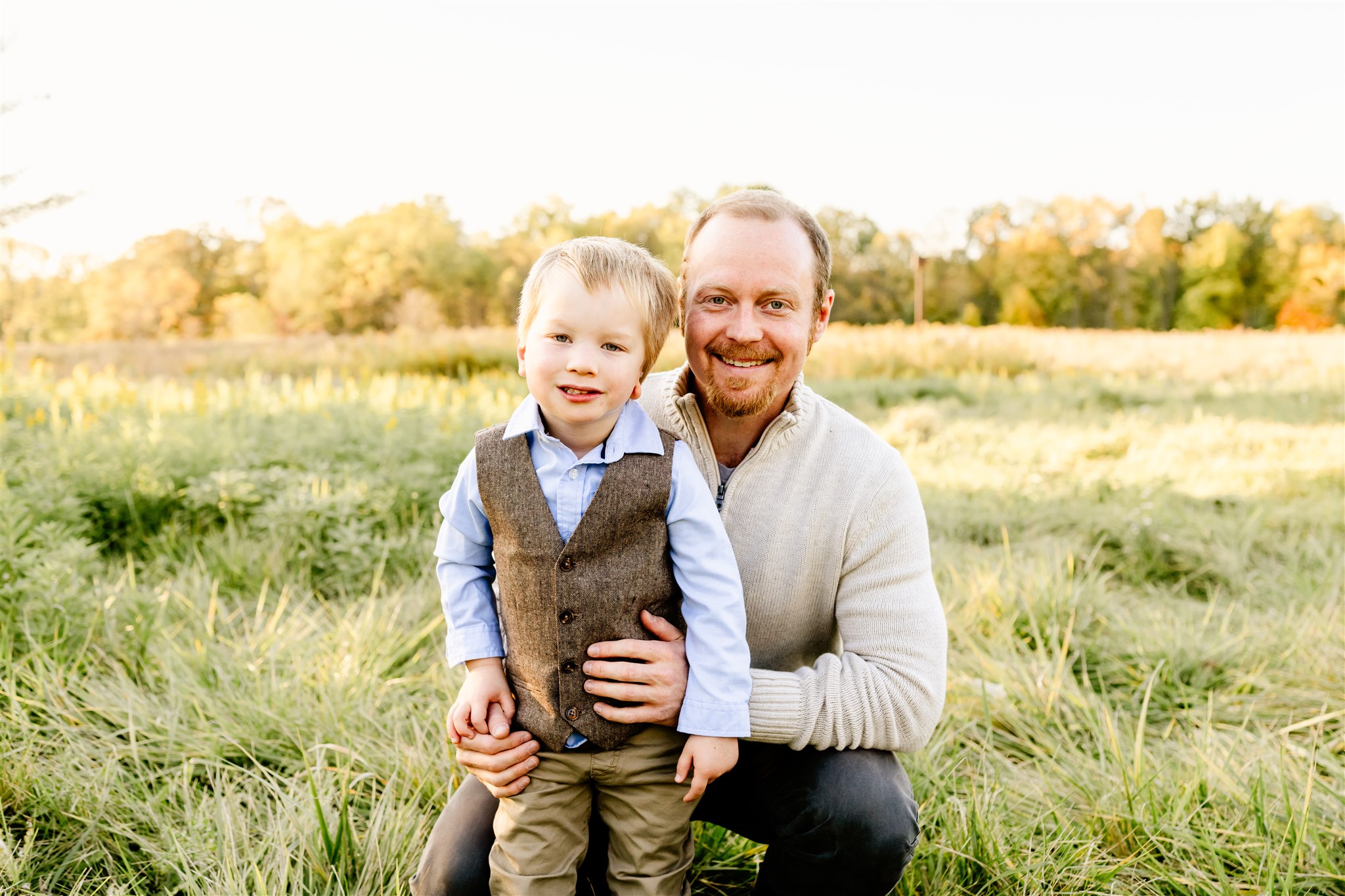 A happy father kneels in some tall grass in a park with his toddler son in front of him
