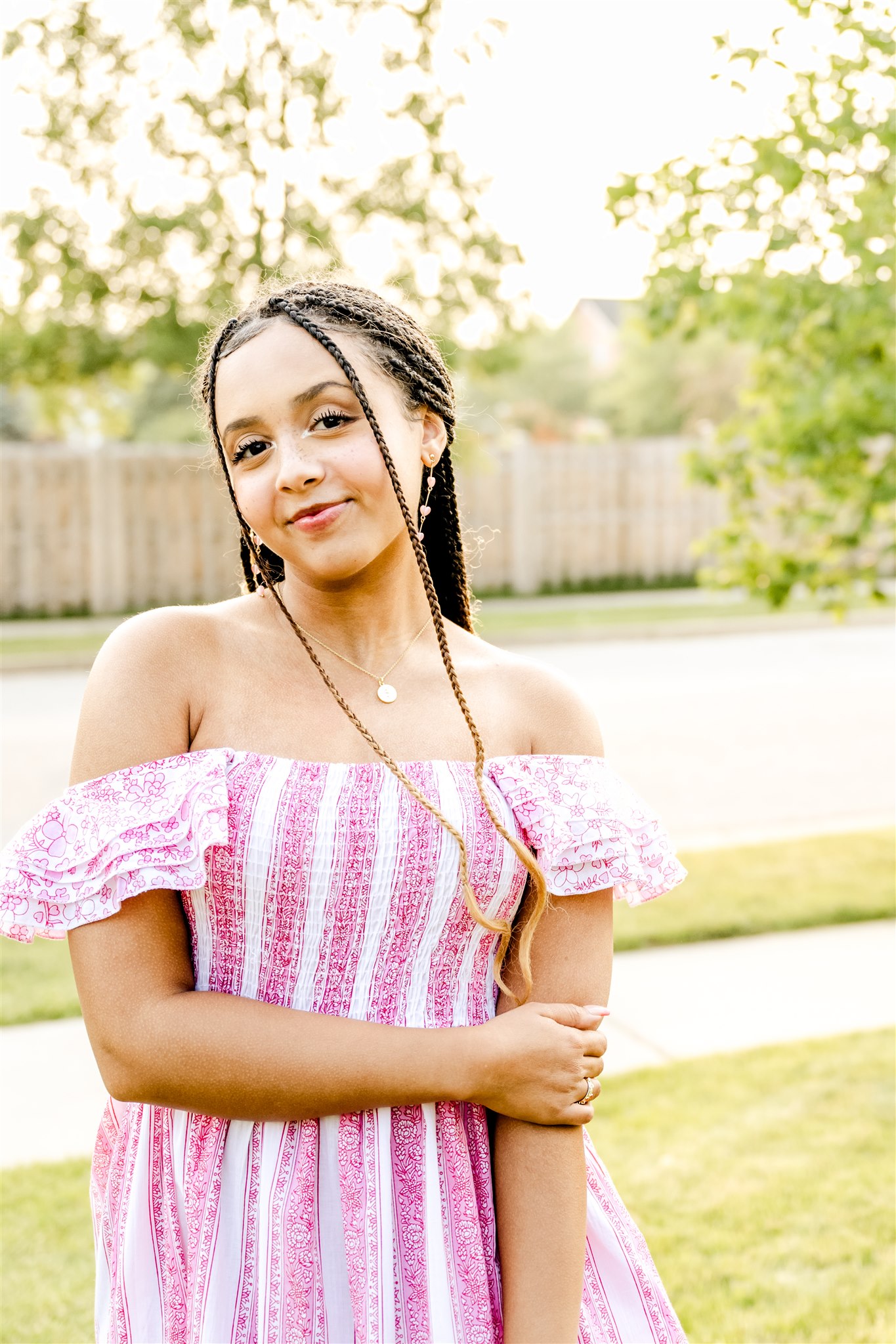 A high school senior stands in her yard in a pink striped dress holding one arm