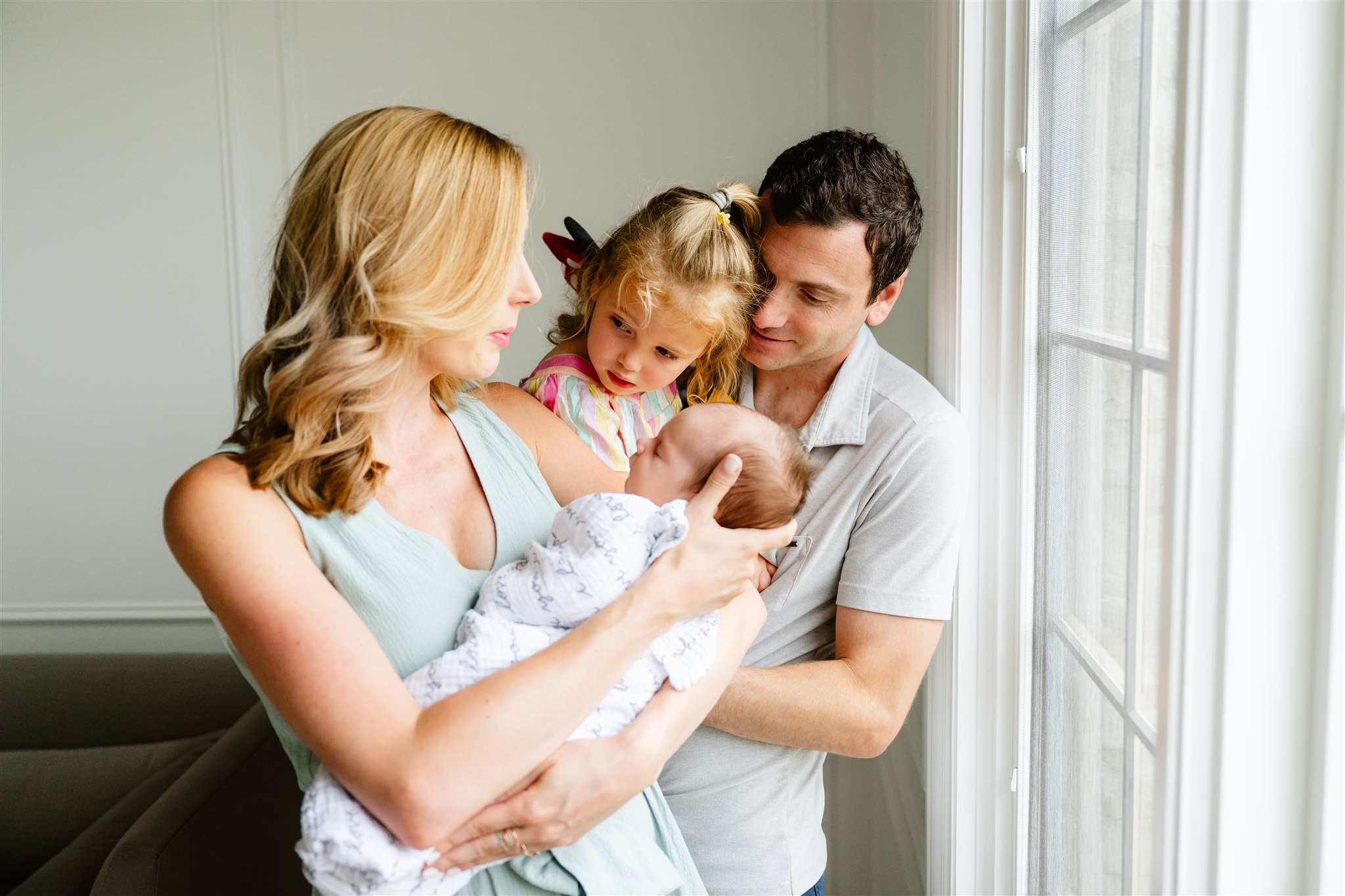 A mom and dad stand in a window holding their newborn baby and toddler daughter in their arms