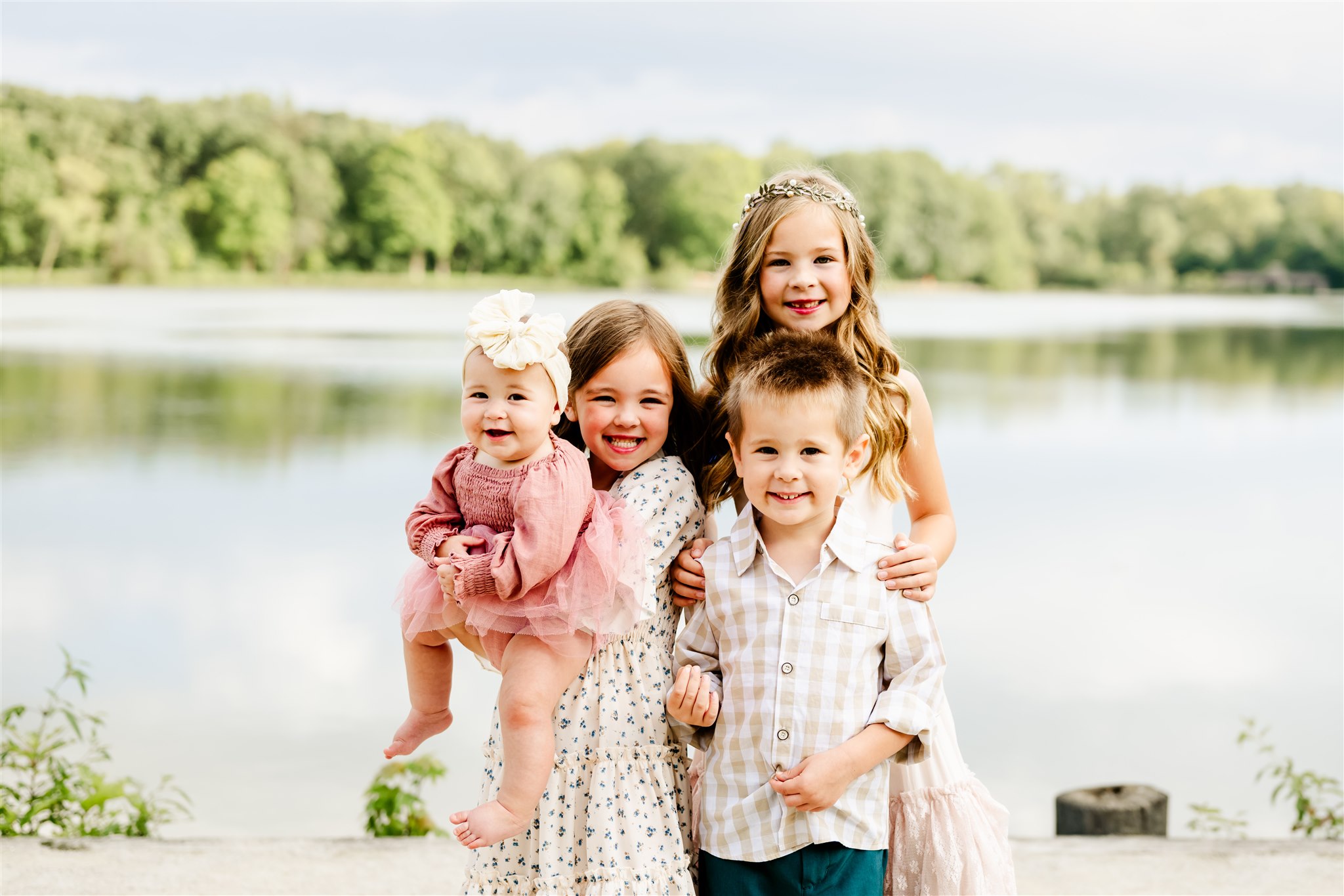Three happy toddler siblings stand by a park pond while one holds baby sister before visiting children's boutiques in Chicago