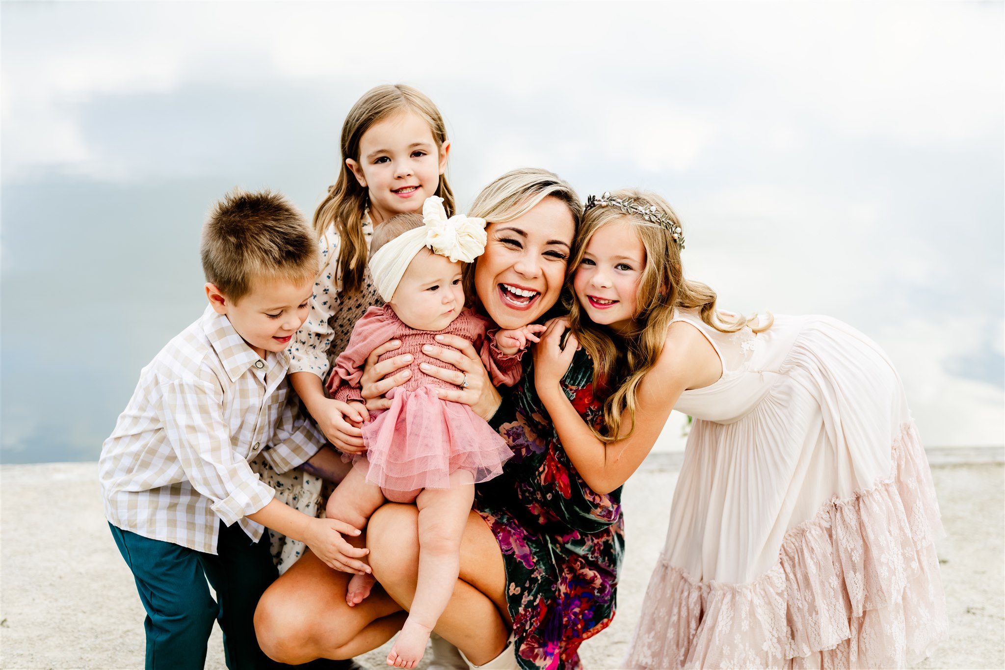 A happy mom is hugged by her three toddlers while holding a baby girl on a beach before visiting children's boutiques in Chicago