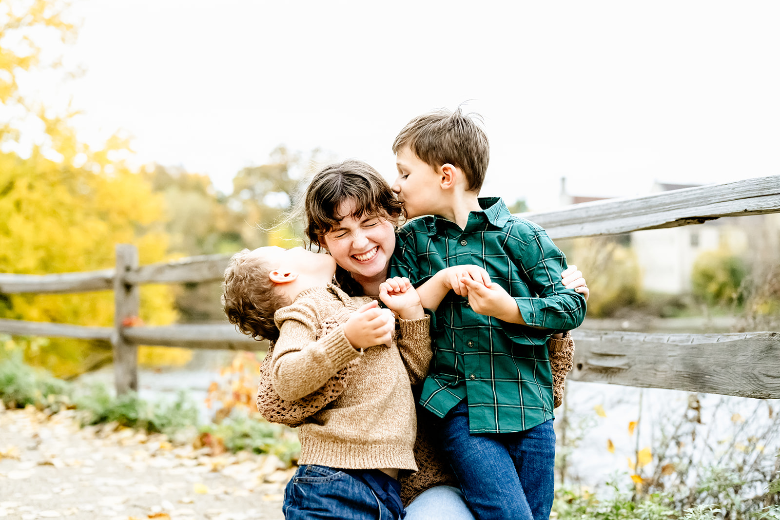 Two toddler boys in jeans kiss their big sister on the cheeks by a park pond after attending christian summer camps in Illinois