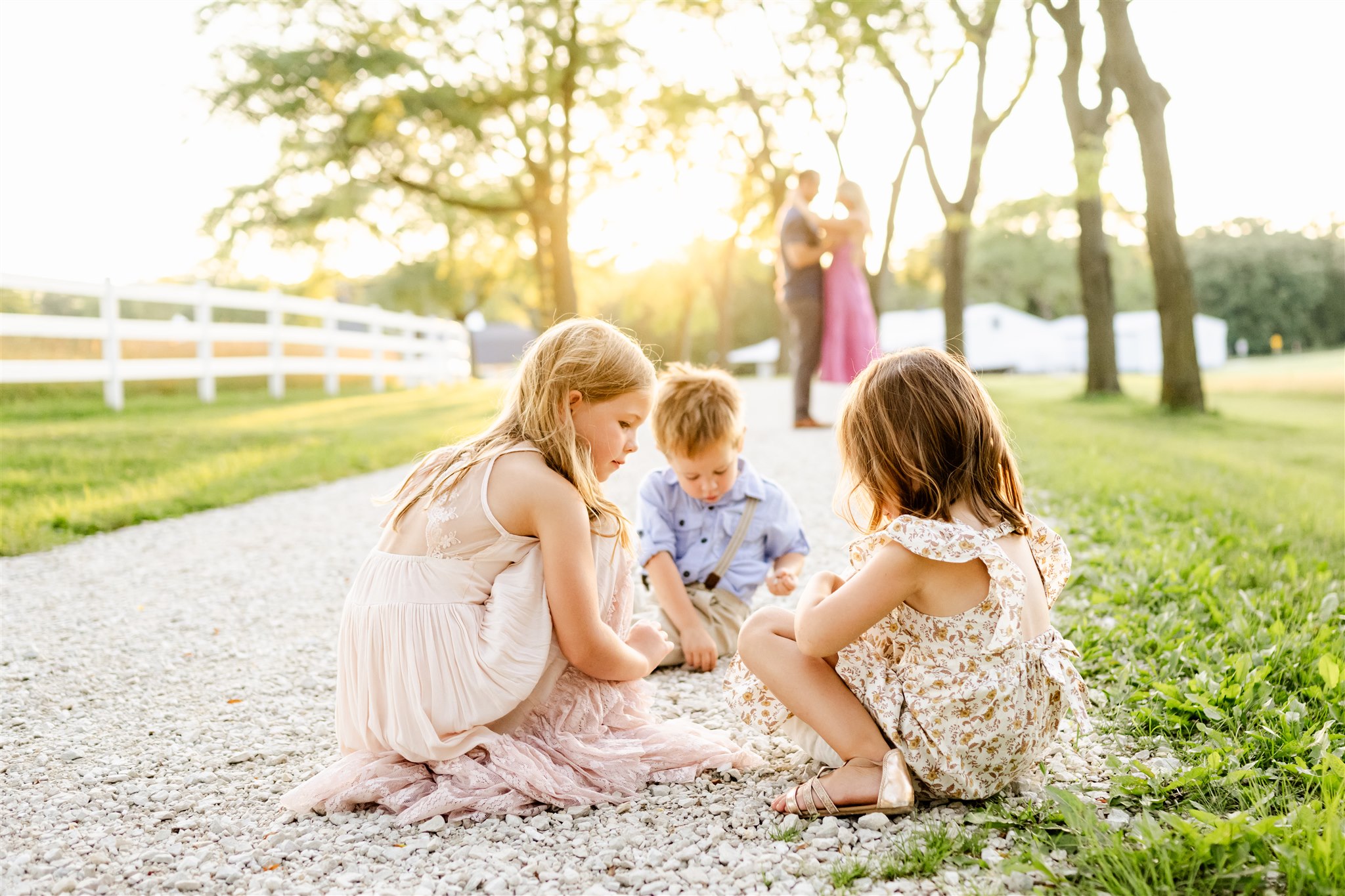 Three young siblings play in a park path as mom and dad snuggle behind them at sunset