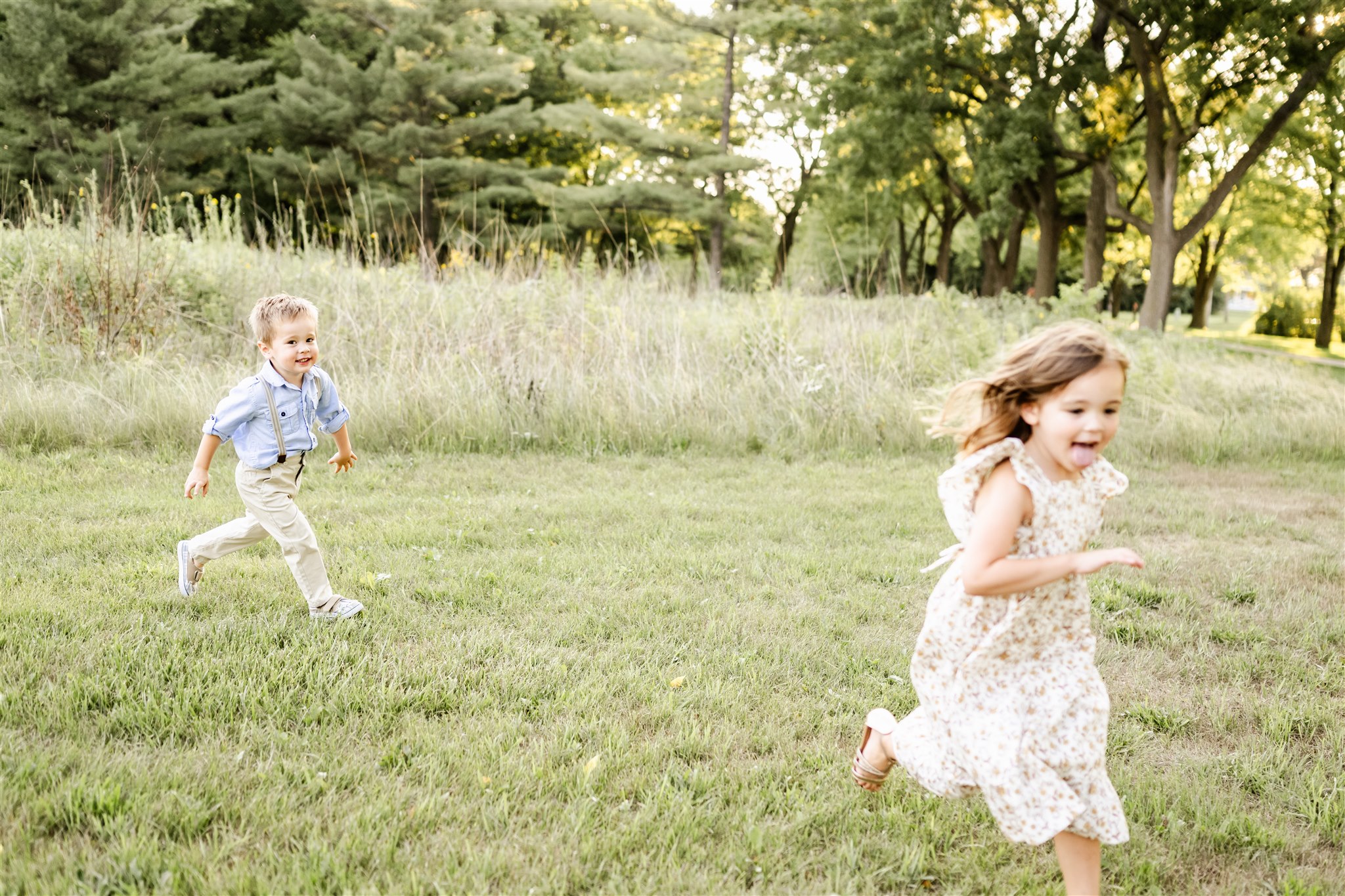 A toddler boy in suspenders and a blue shirt chases his sister in a dress in a park after some kids swim lessons in Chicago