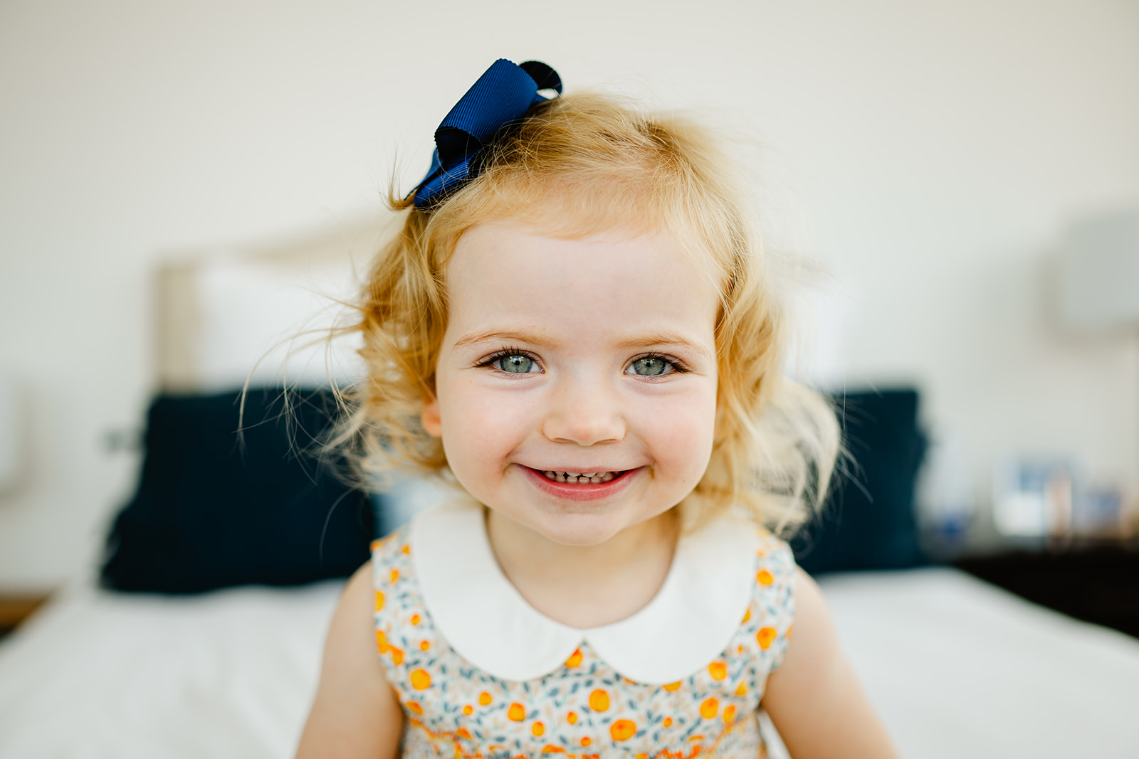 A happy toddler girl in a yellow dress smiles big on a bed