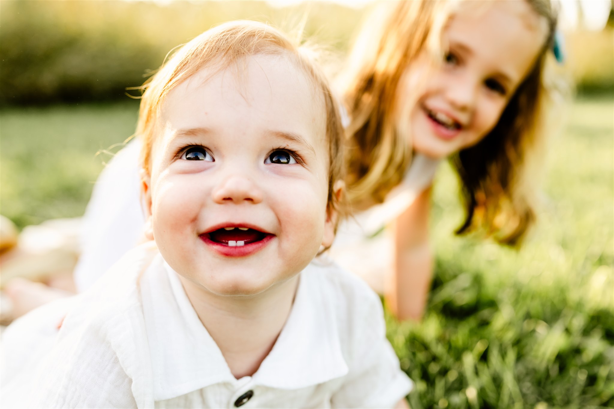 A baby in a white shirt crawls in a park lawn with older sisters in a white dress behind him