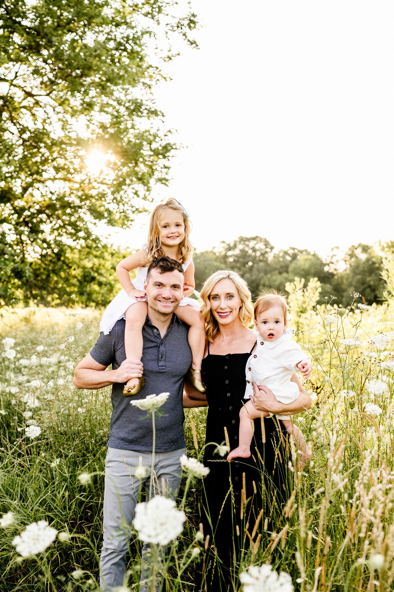 Happy mom and dad stand in a field of tall wildflowers with their baby and toddler girl on their shoulders after some private swim lessons in Naperville