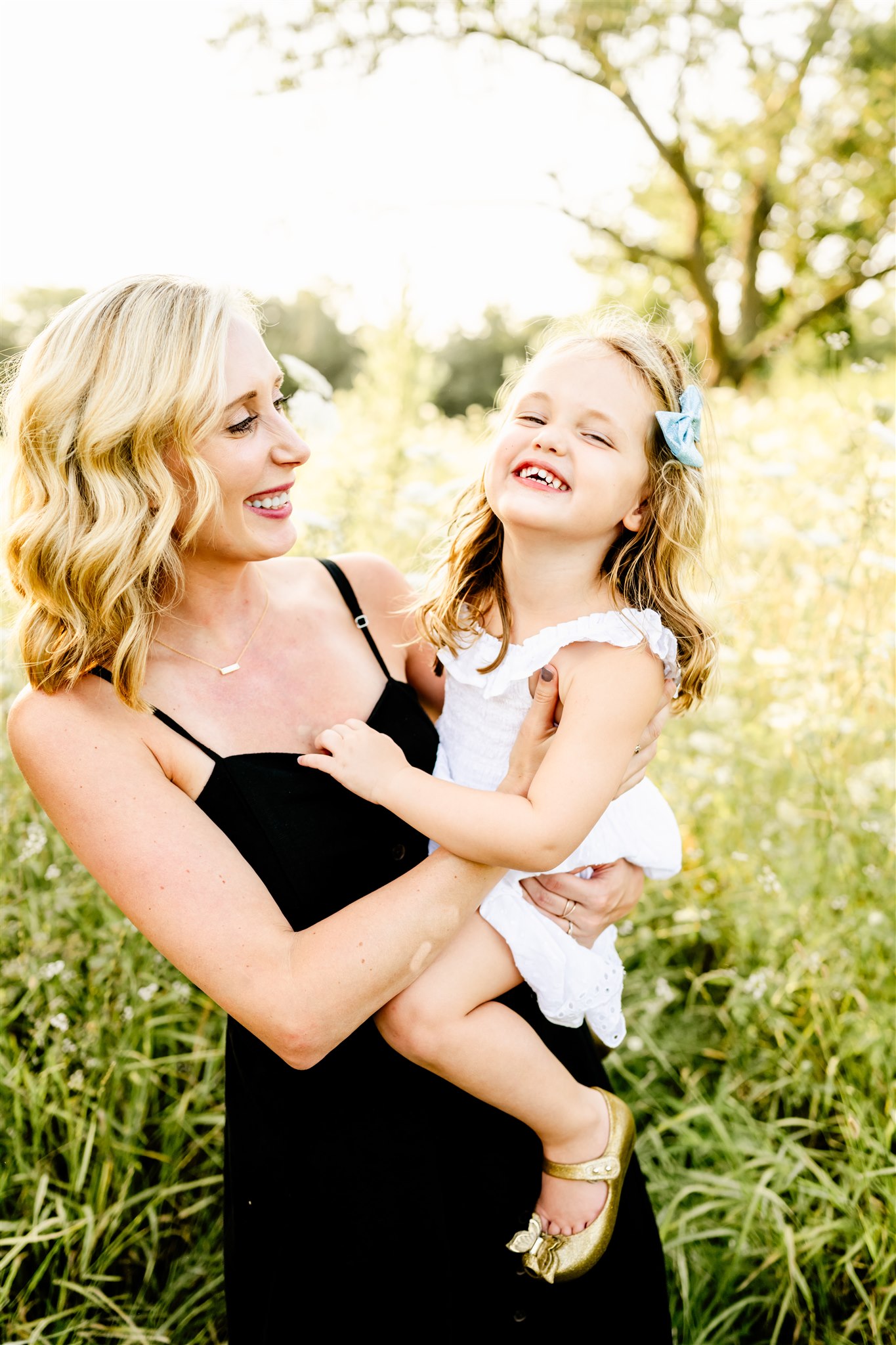 A happy toddler girl in a white dress sits on mom's hip while they laugh and explore a park after some private swim lessons in Naperville