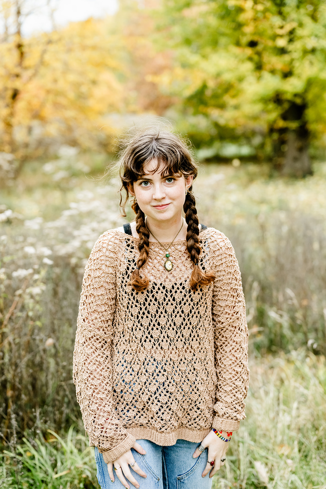 A teenage girl in a brown blouse and jeans stands in a park by some wildflowers at sunset