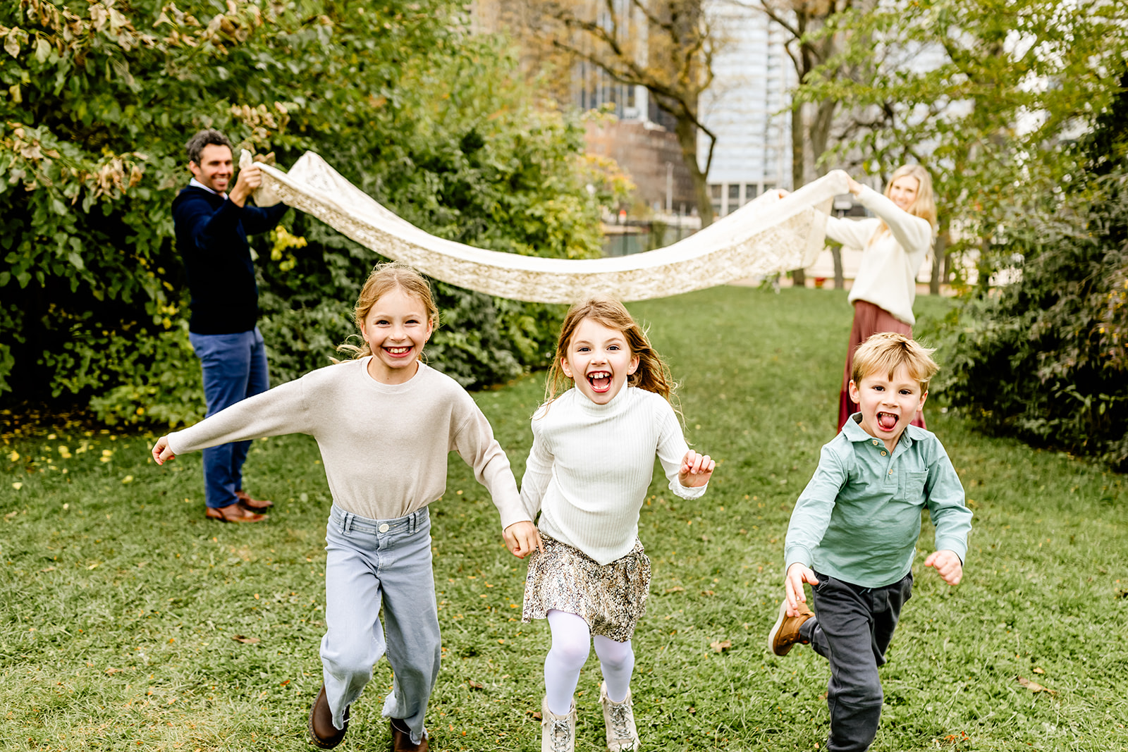 A young boy runs in a park lawn with his two sisters under a blanket held by mom and dad