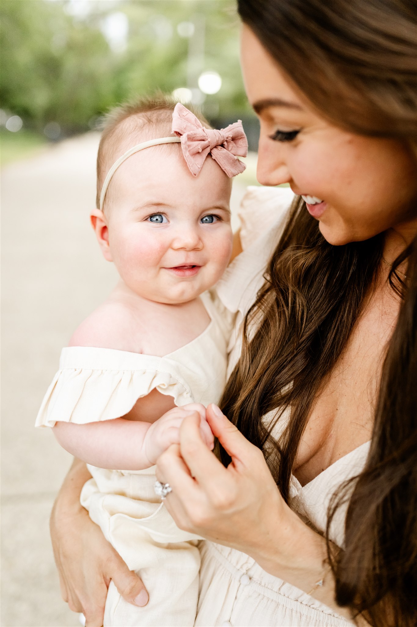 A baby girl in a white dress smiles while sitting in mom's arms in a park sidewalk
