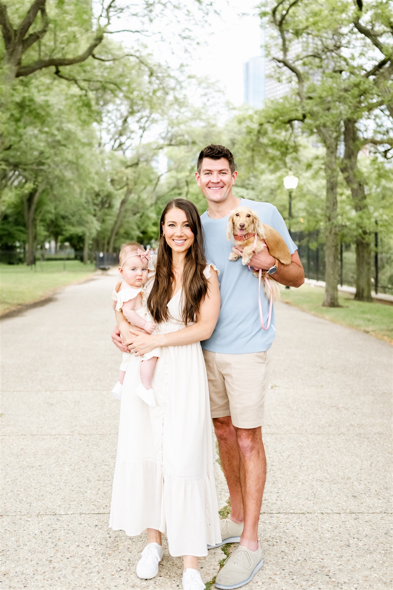 Happy mom and dad stand in a park sidewalk with a newborn and tiny dog after visiting hair salons in Naperville