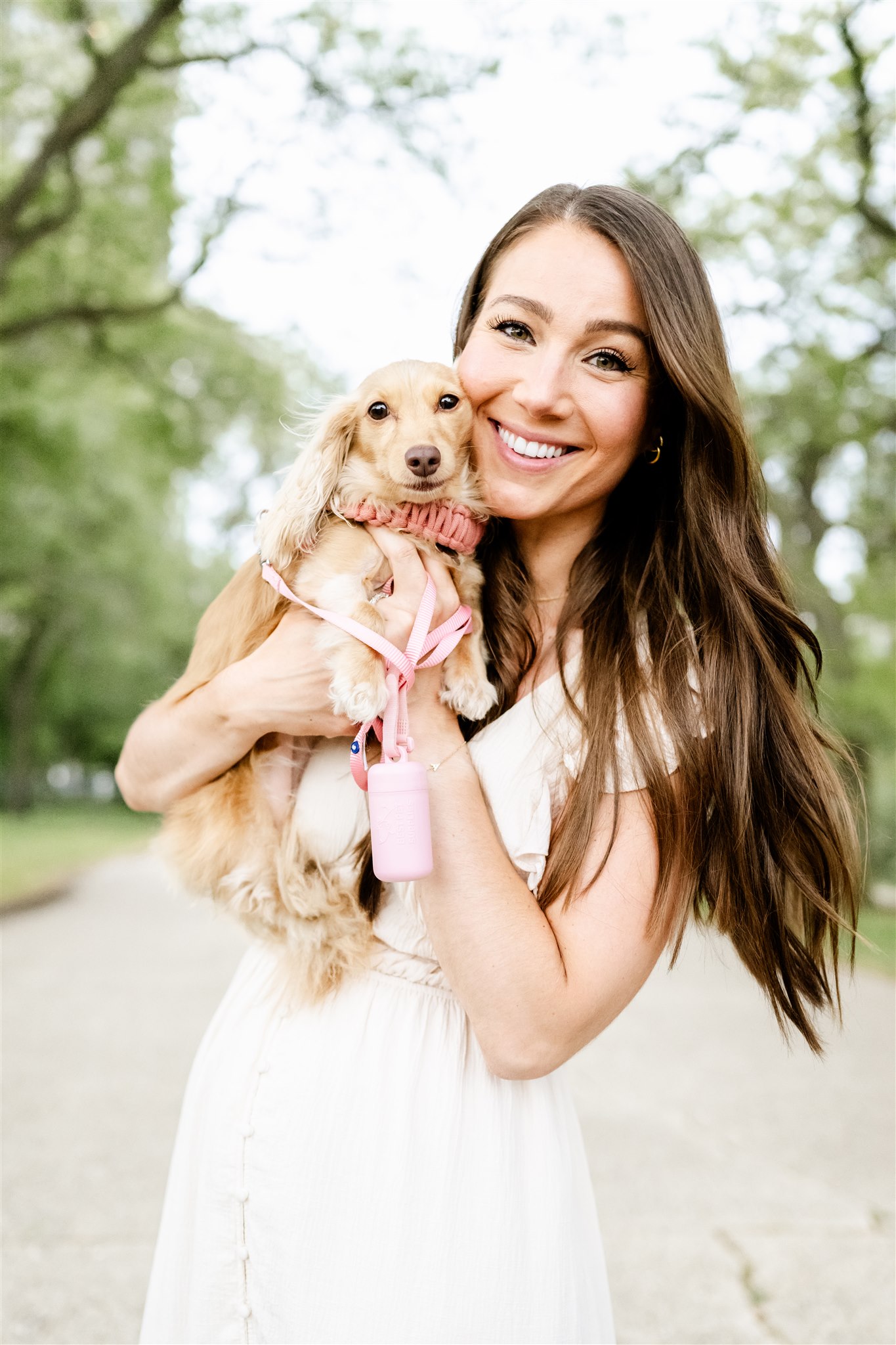 A happy woman in a white dress snuggles a tiny dog in a park after visiting hair salons in Naperville