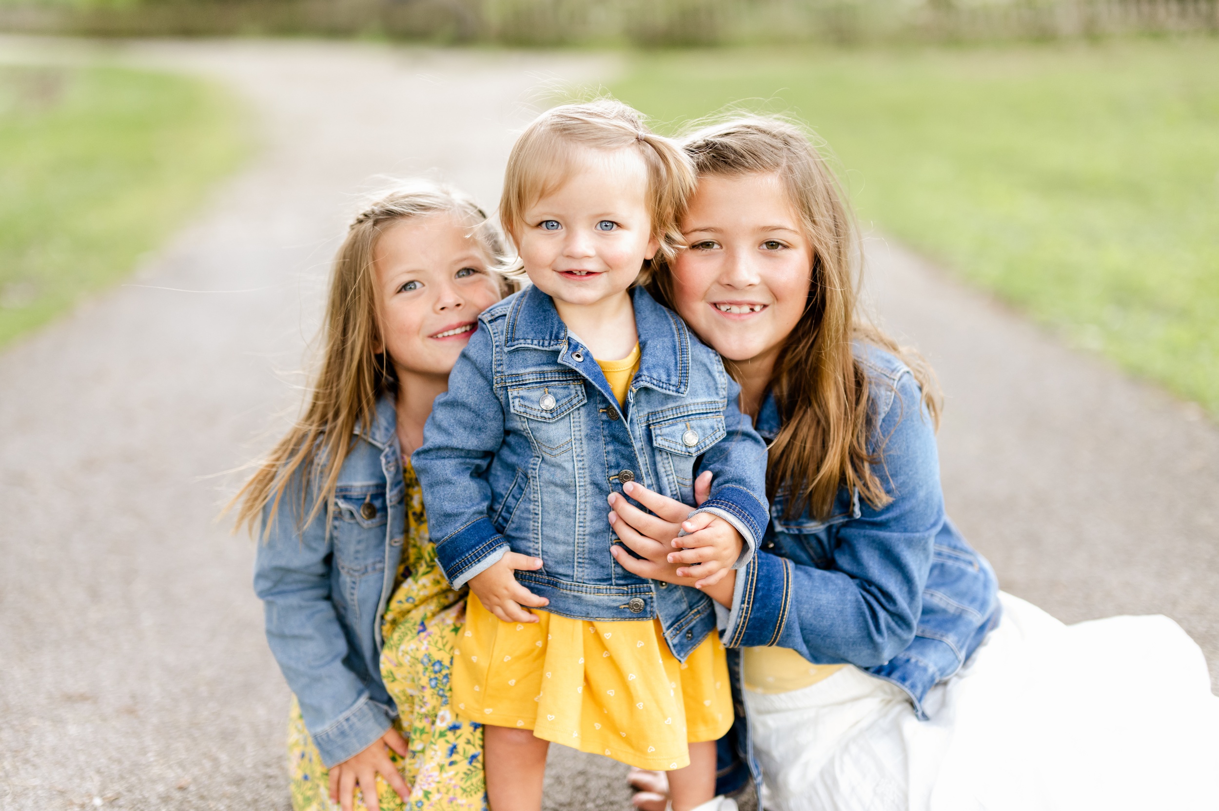 Three toddler sisters in denim jackets and yellow dresses hug and smile in a park sidewalk
