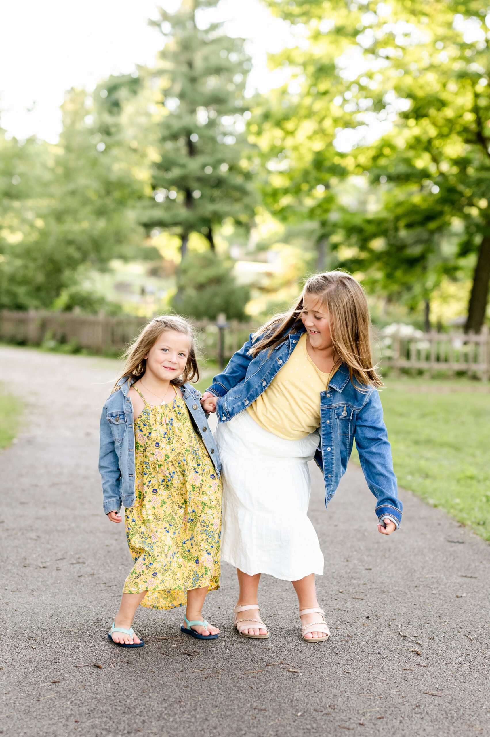 Happy toddler sisters in yellow and denim bump hips in a park sidewalk while playing after montessori schools in naperville