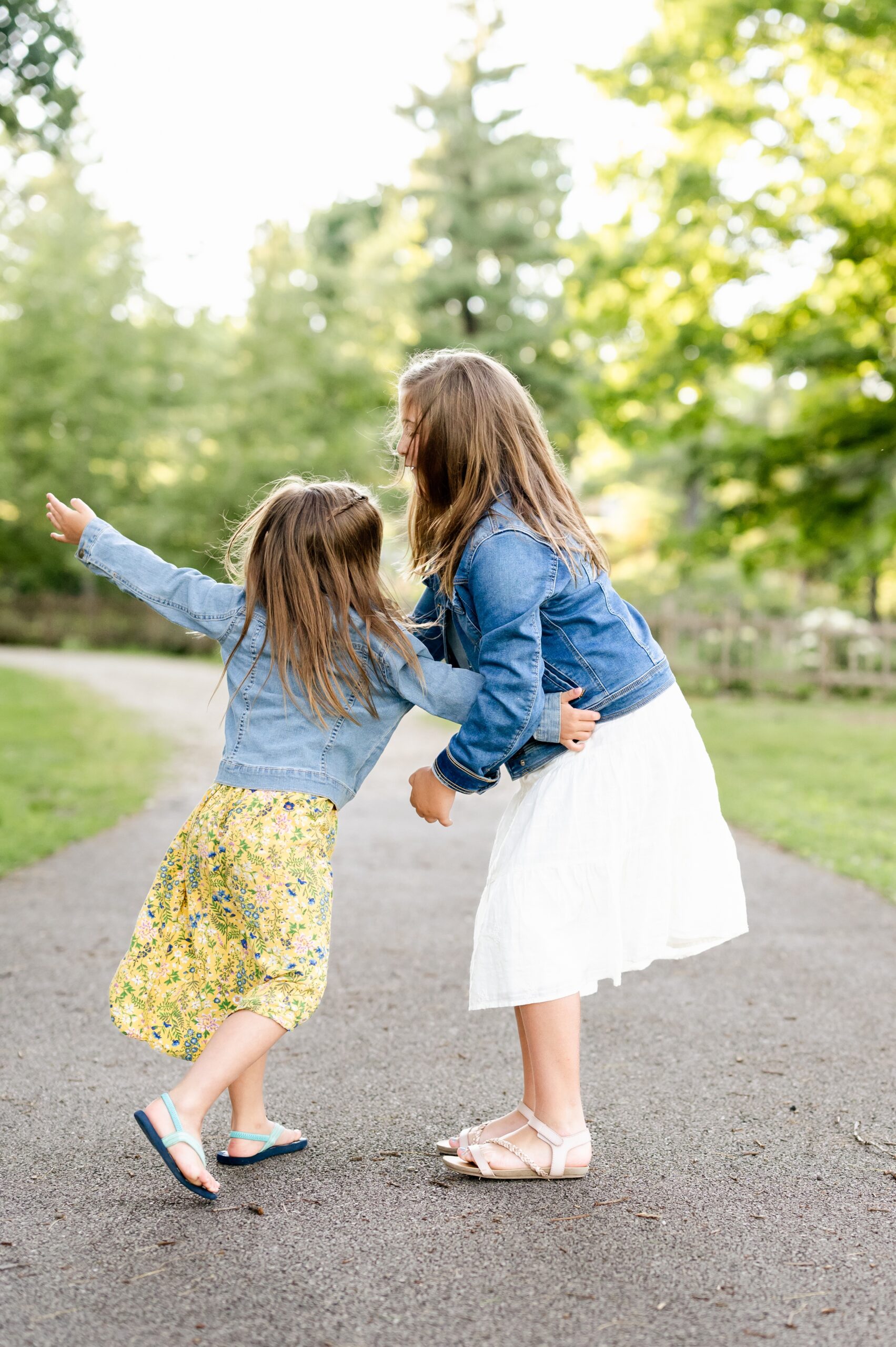 Toddler sisters in yellow and white dresses play and hug in a park sidewalk after leaving montessori schools in naperville