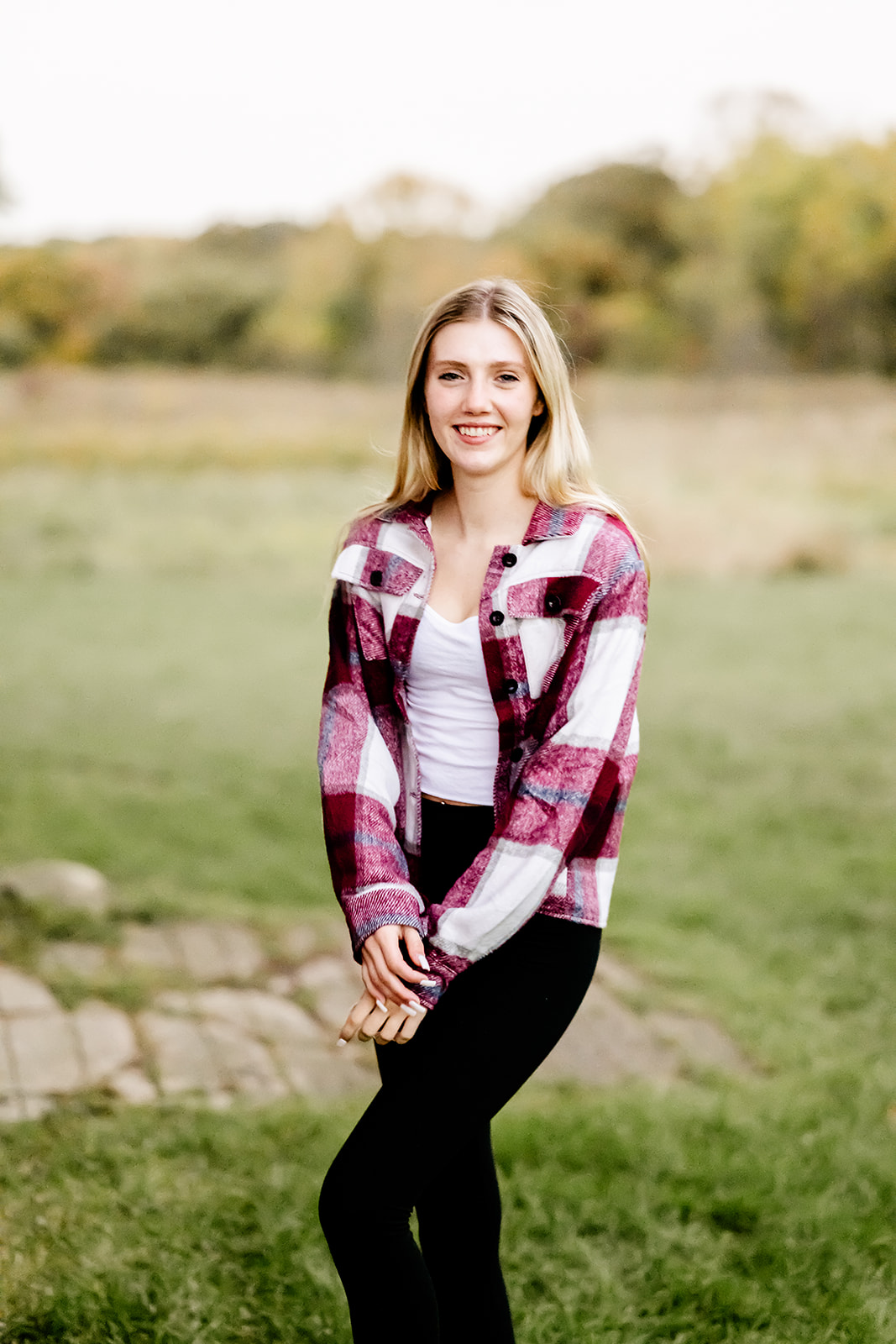 A high school senior in black pants and red flannel shirt smiles while standing in a park at sunset