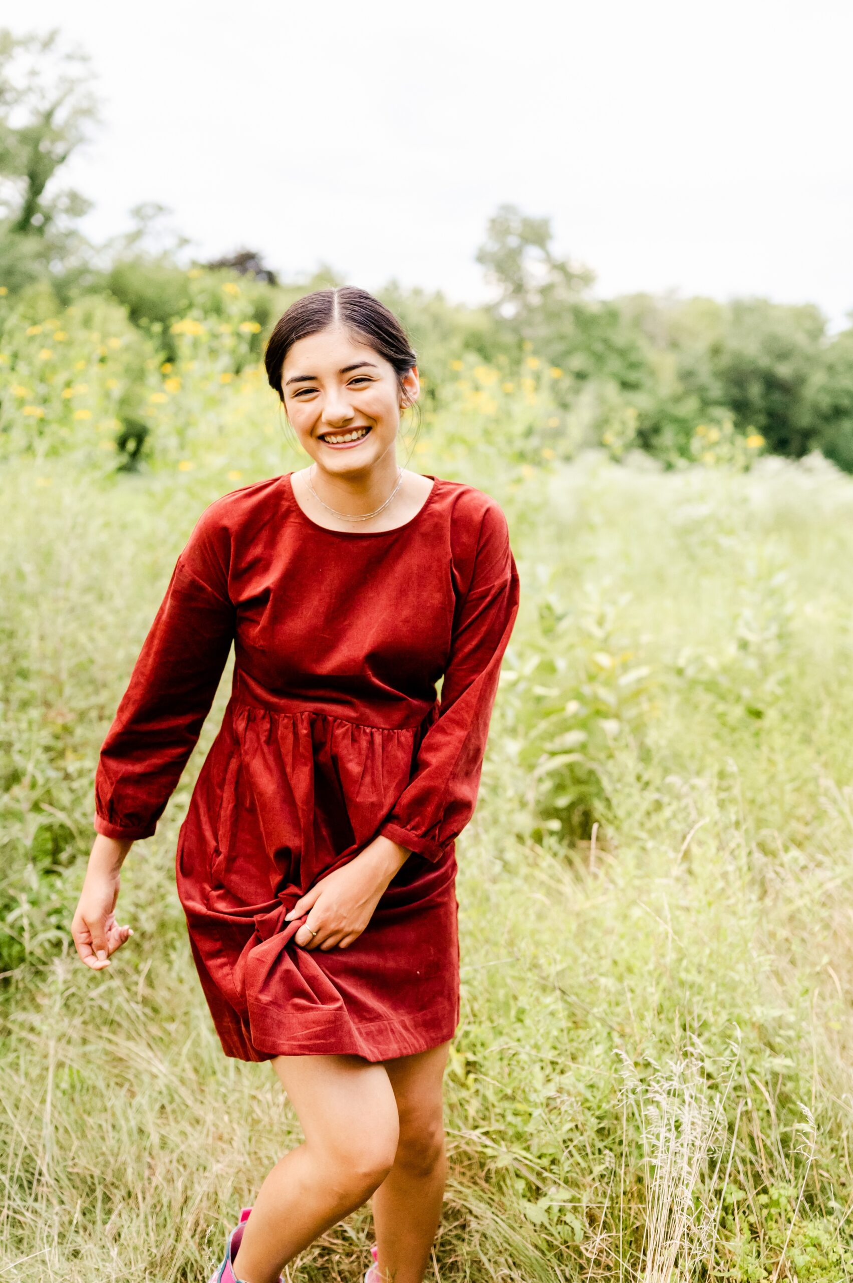 A happy high school senior laughs while playing in tall grass at sunset in a red dress after visiting nail salons in Naperville