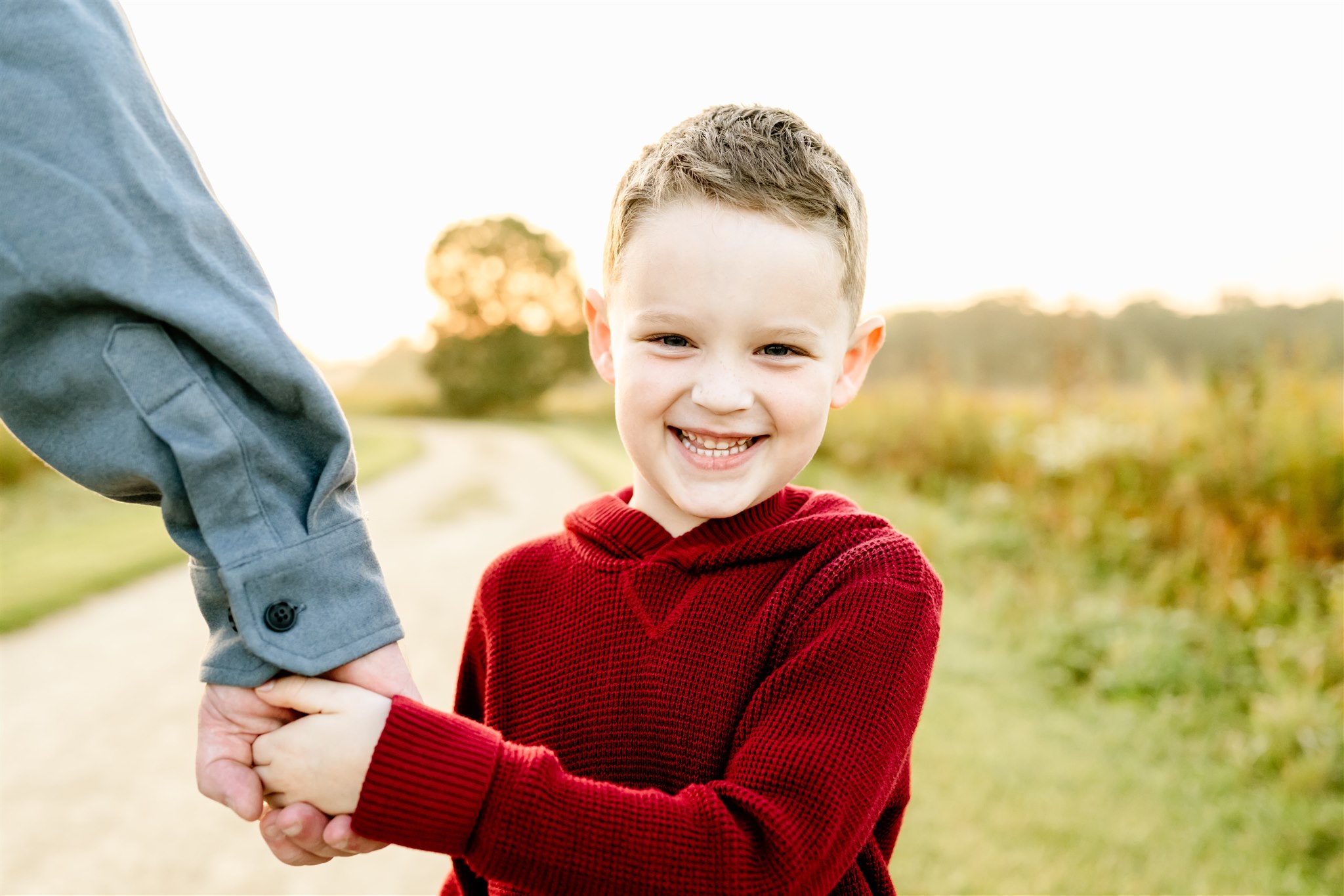 A happy toddler in a red sweater holds dad's hand in a park trail while giggling