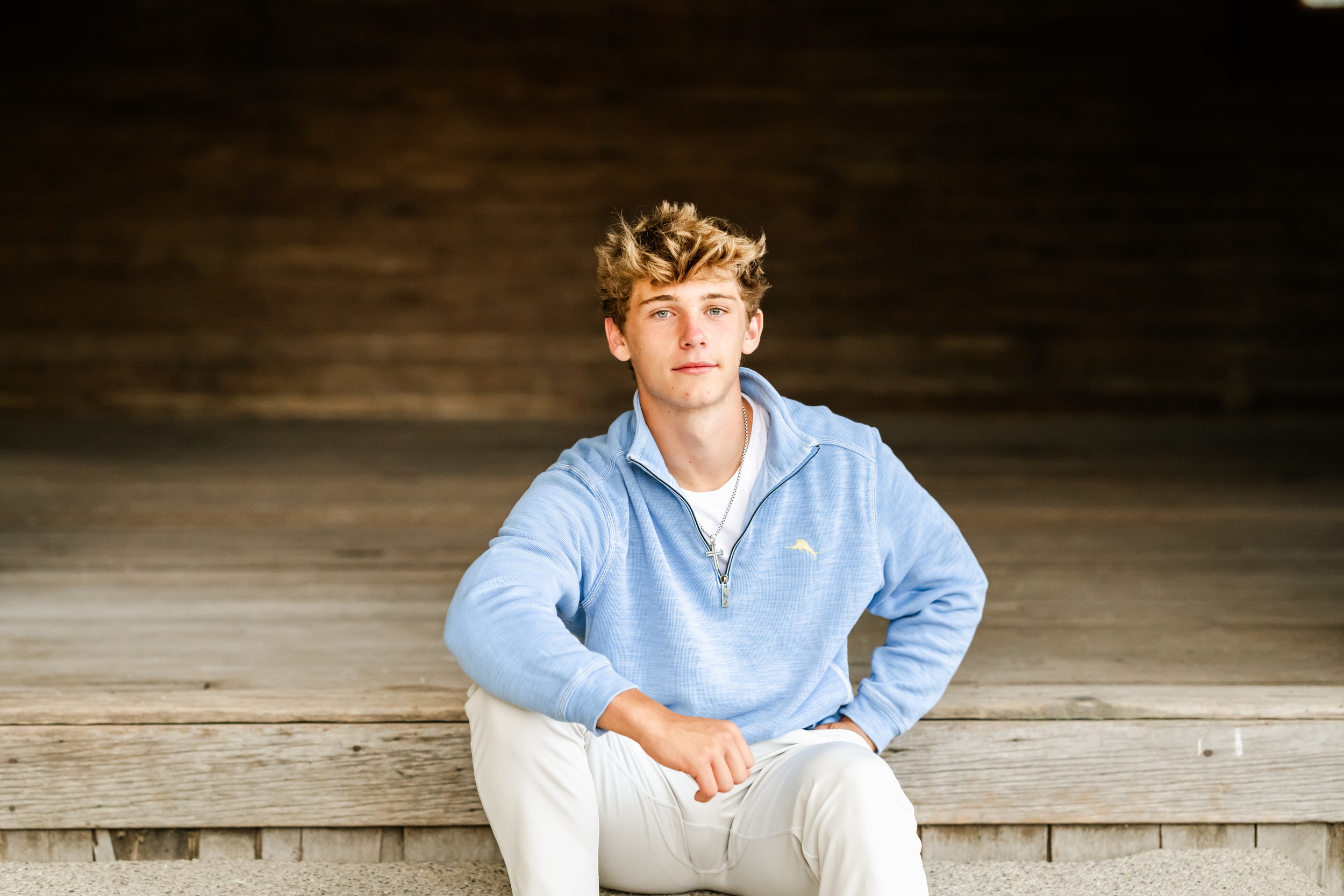 A high school senior sits on steps of a rustic building in a blue sweater and light pants
