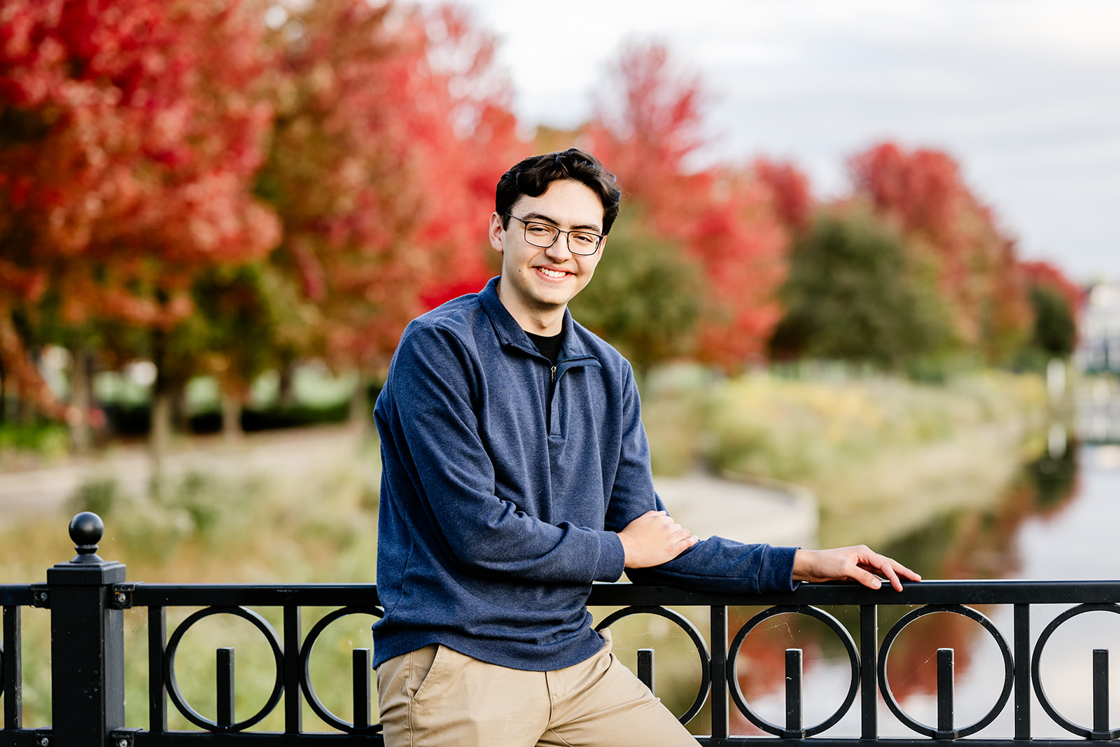 A teenager in a blue sweater leans on an iron fence on a pedestrian bridge over a canal in fall