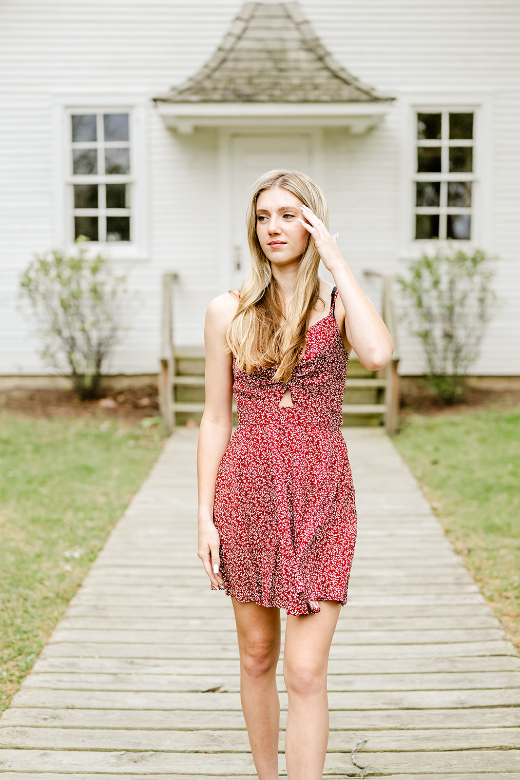 A woman in a res dress walks on a park boardwalk by a historic building with a hand in her hair