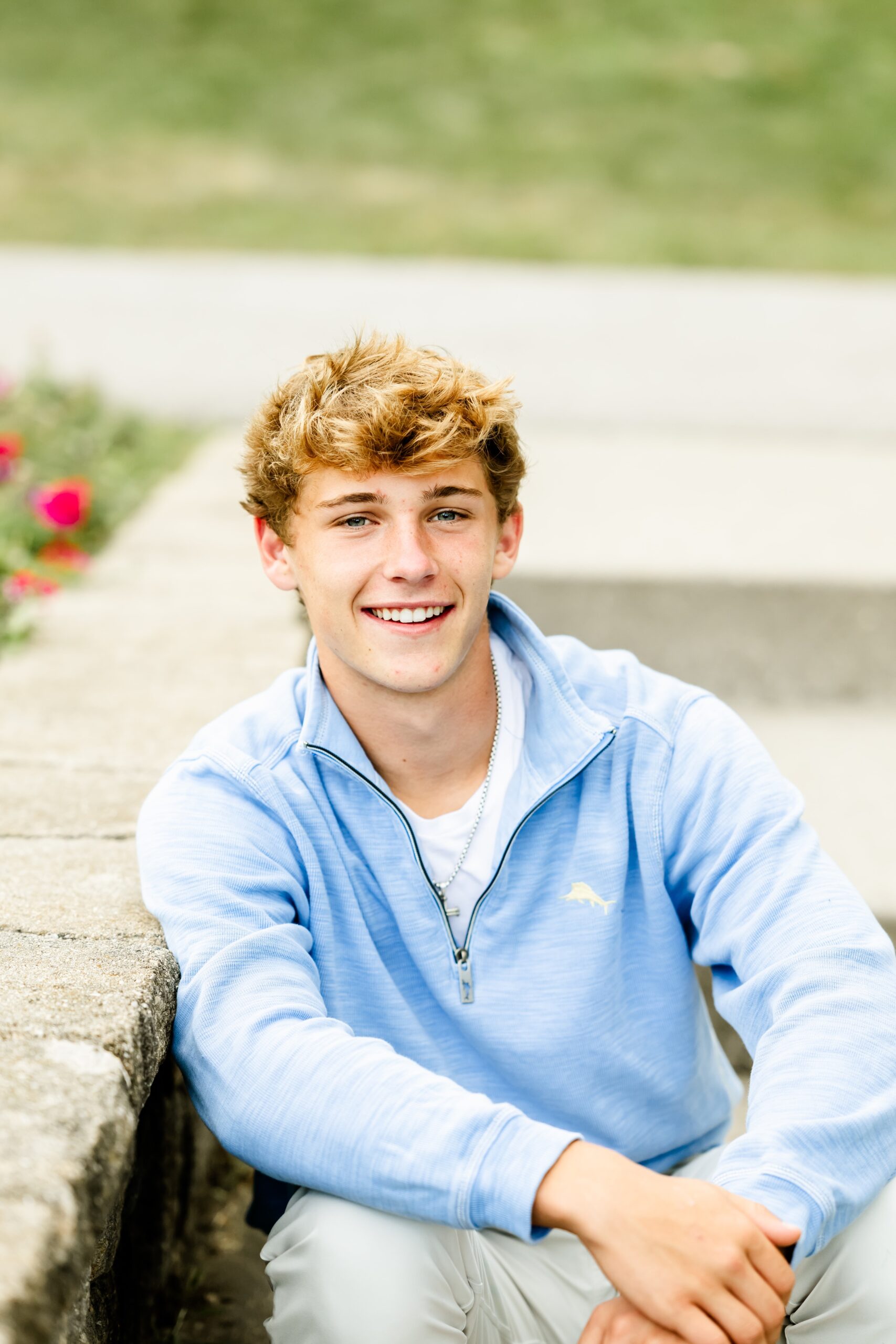 A smiling high school senior in a blue sweater leaning on a garden wall while sitting on steps
