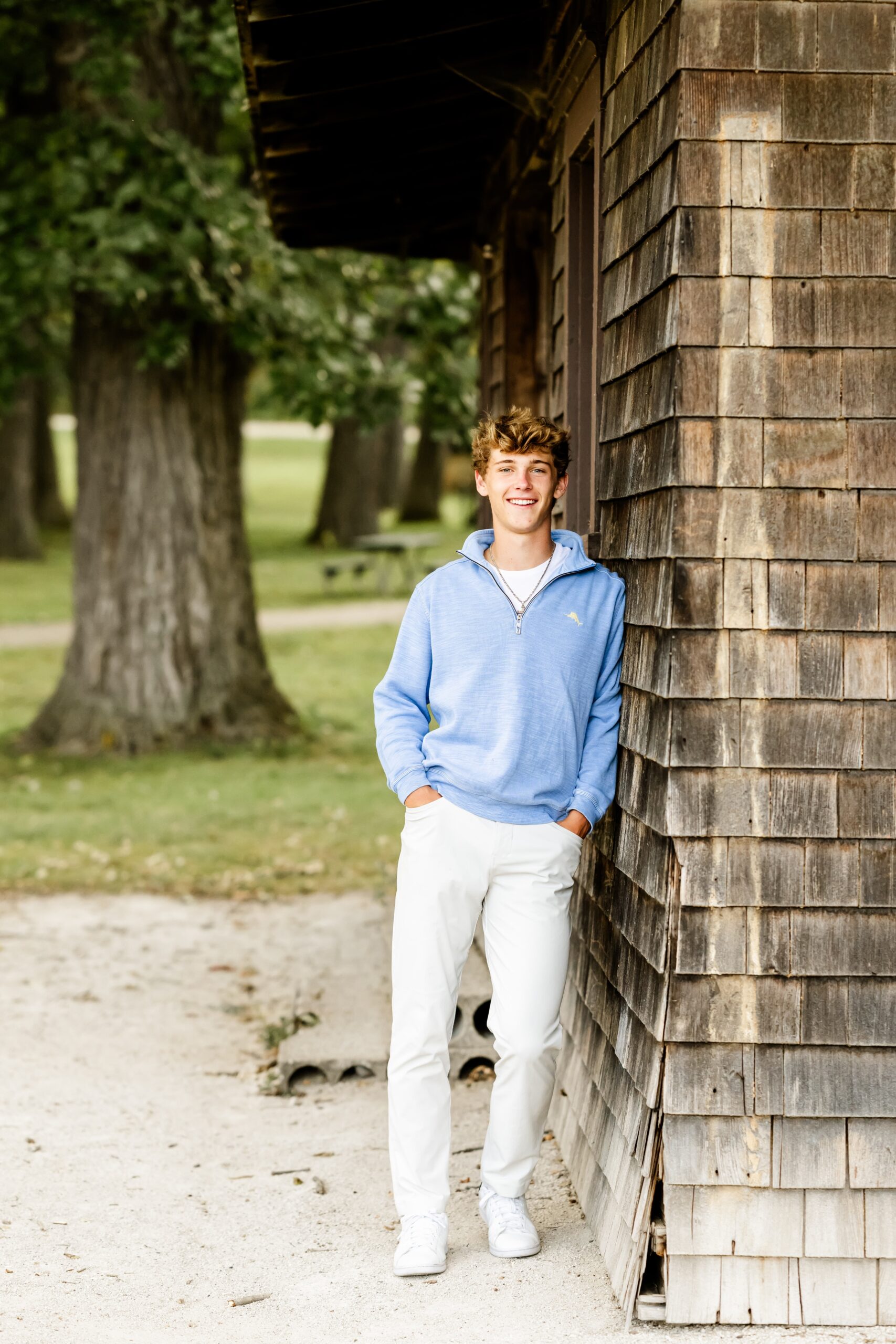 A happy teenager smiles in a blue sweater while leaning on a park building in white pants after using SAT prep in Naperville