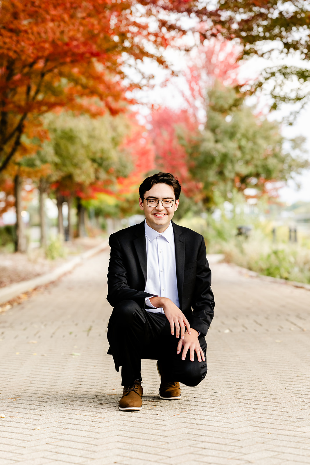 A high school senior in a black and white suit kneels in a park sidewalk after using tutors in Naperville