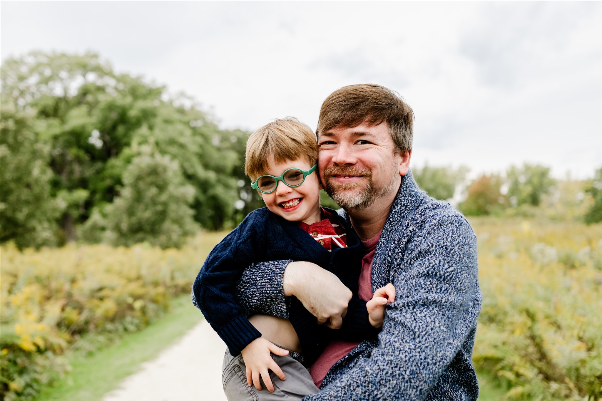 A smiling father in a sweater hugs his toddler son in a park path