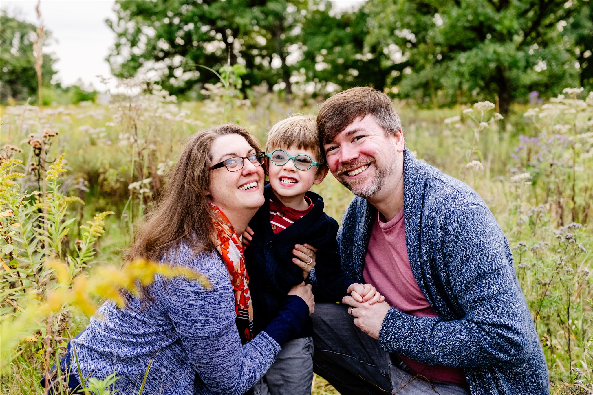 Happy mom and dad in sweaters kneel in a garden witht heir smiling toddler son in a dark sweater and glasses after visiting Children's Museums in Chicago